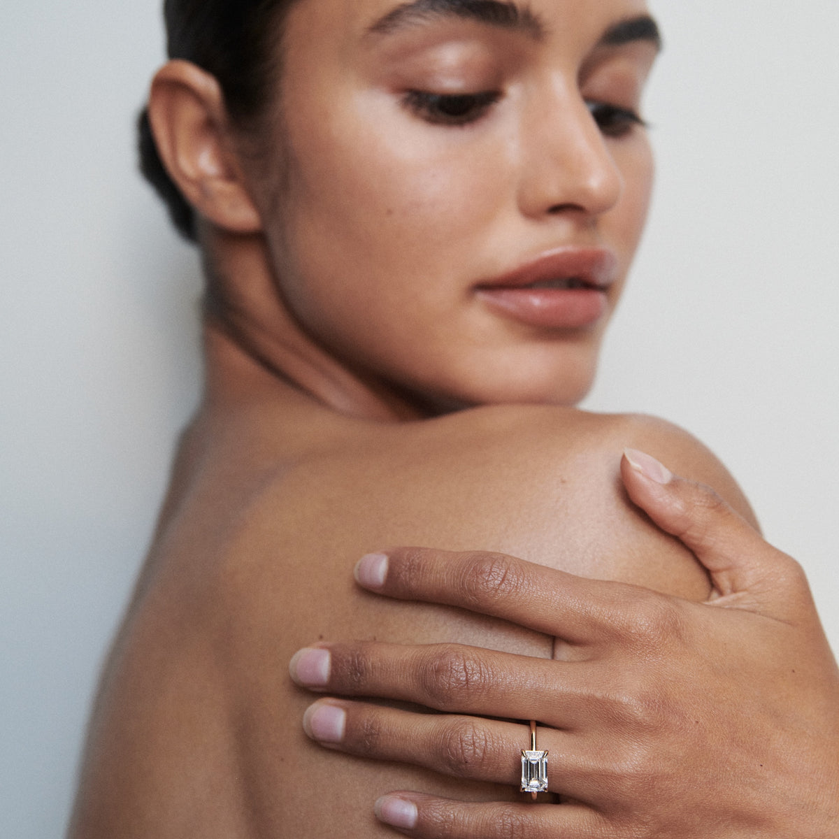 Woman showcasing a luxurious, emerald-cut diamond ring on her finger, against her bare shoulder. Elegant and minimalist jewellery display.