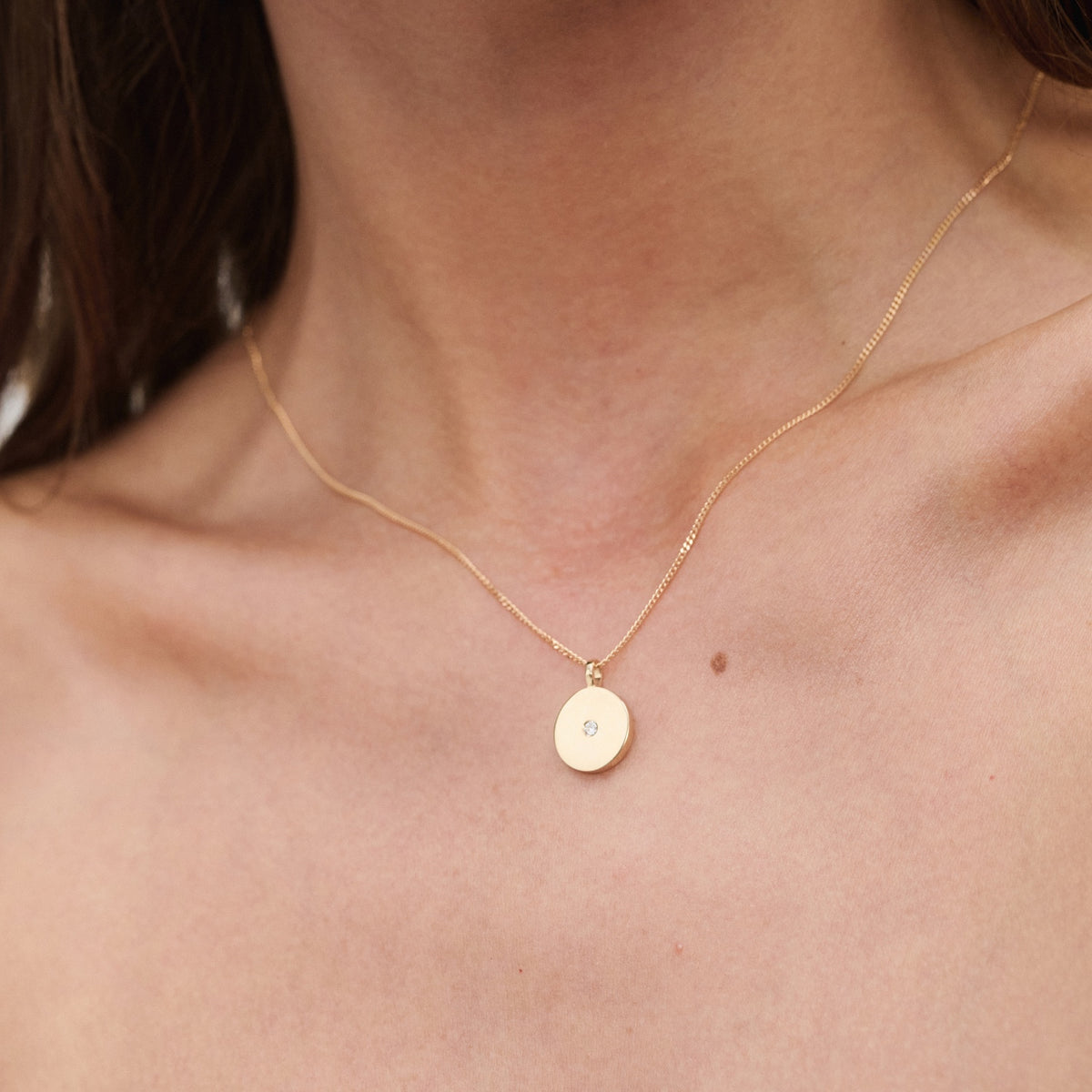 Gold necklace with a round pendant featuring a small diamond, worn around a woman's neck; minimalist luxury jewellery.