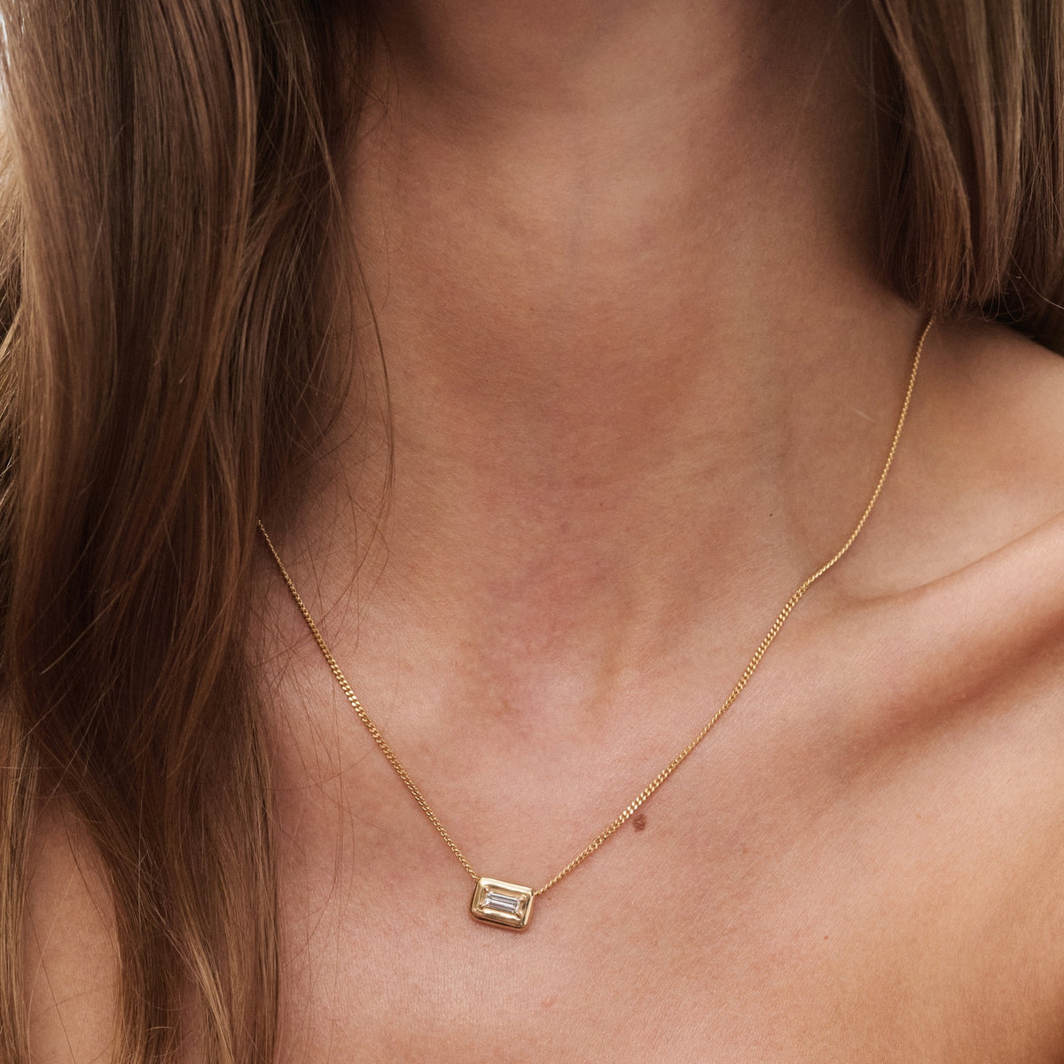 Woman wearing a gold necklace with an emerald-cut pendant. The jewellery rests elegantly on her collarbone, showcasing minimalist luxury.