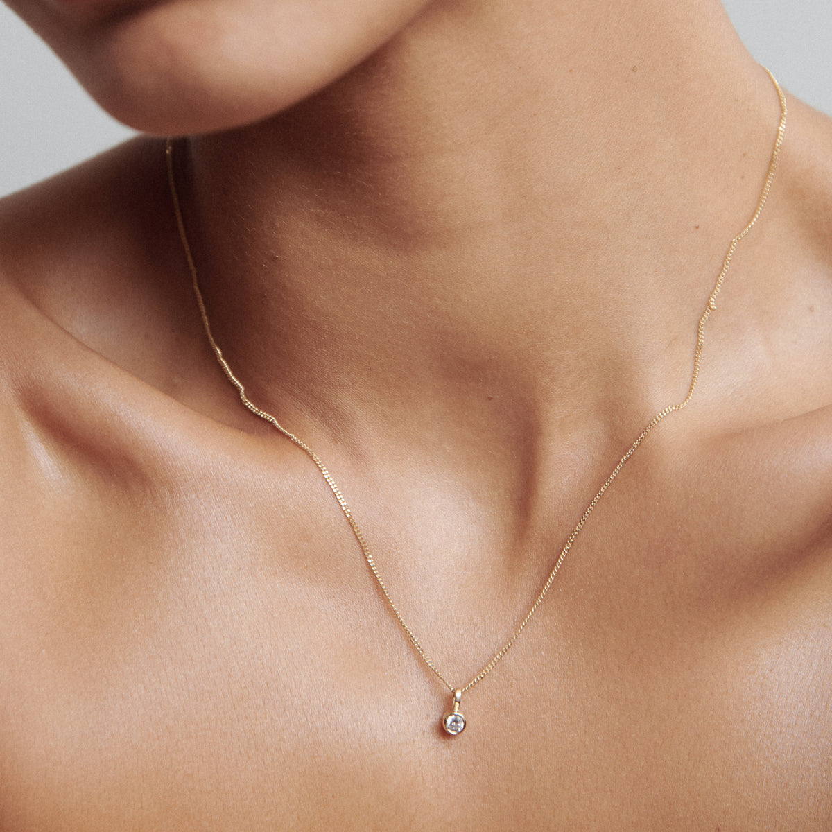 Elegant gold pendant necklace with a small diamond, worn around a person's neck. Luxurious and minimalist jewellery design.