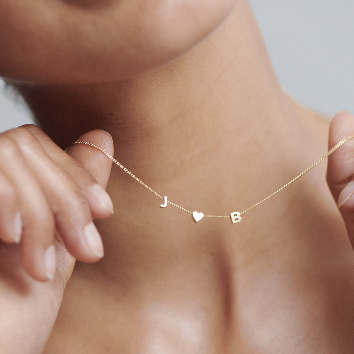 Woman holding delicate gold necklace with letter charms "J", heart, and "B". Perfect luxury jewellery for personal style.