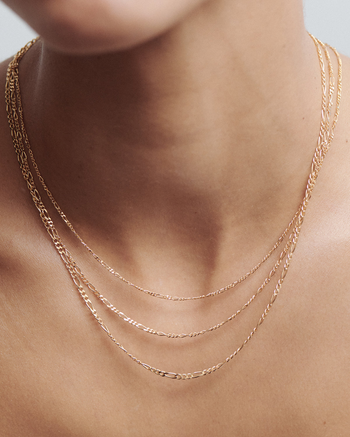 Close-up of a person wearing three delicate layered gold chains. The fine chain necklaces rest elegantly on a bare neck, showcasing luxury jewellery detail.