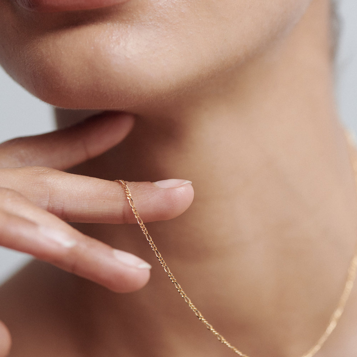 Close-up of a woman's neck and hand, featuring a delicate gold chain necklace draped over her fingers.[text overlay: Small Figaro]