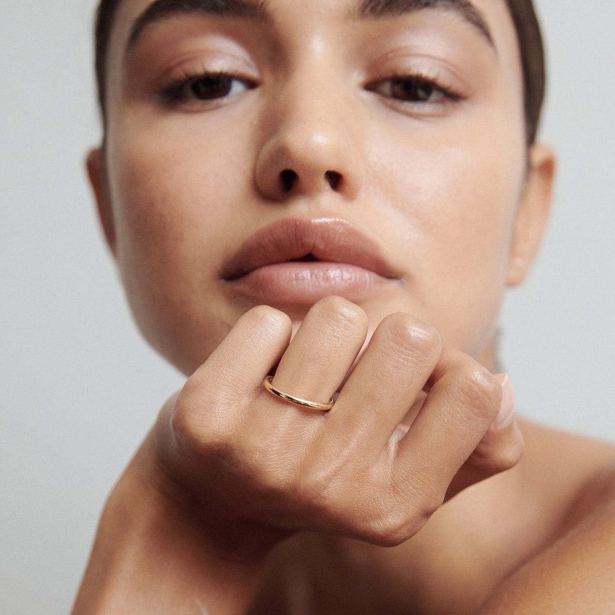 Woman with natural makeup gazes forward, resting chin on hand adorned with an elegant gold ring. Minimalist luxury jewellery.