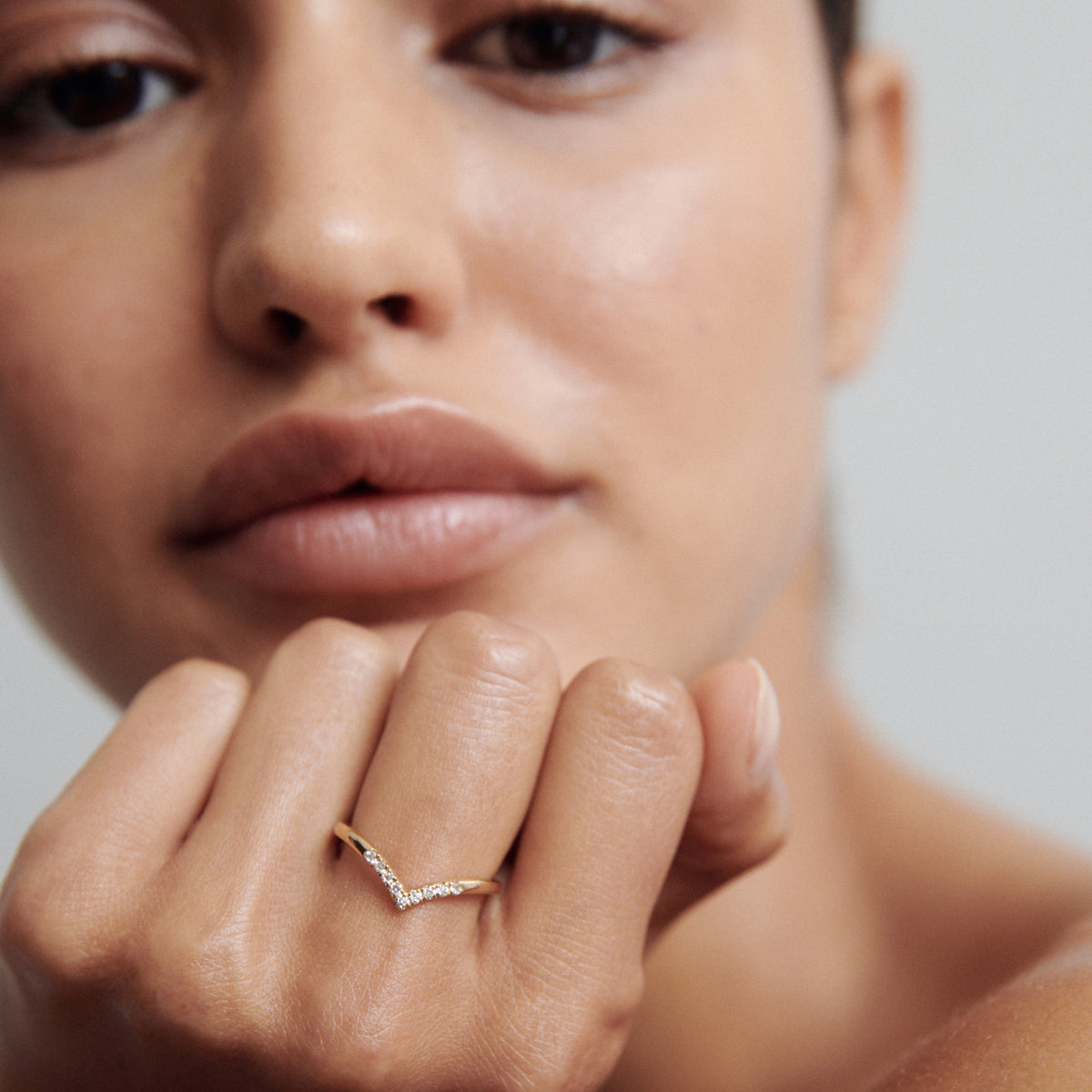 Woman wearing a delicate gold ring with a chevron design, adorned with small diamonds, positioned near her lips.