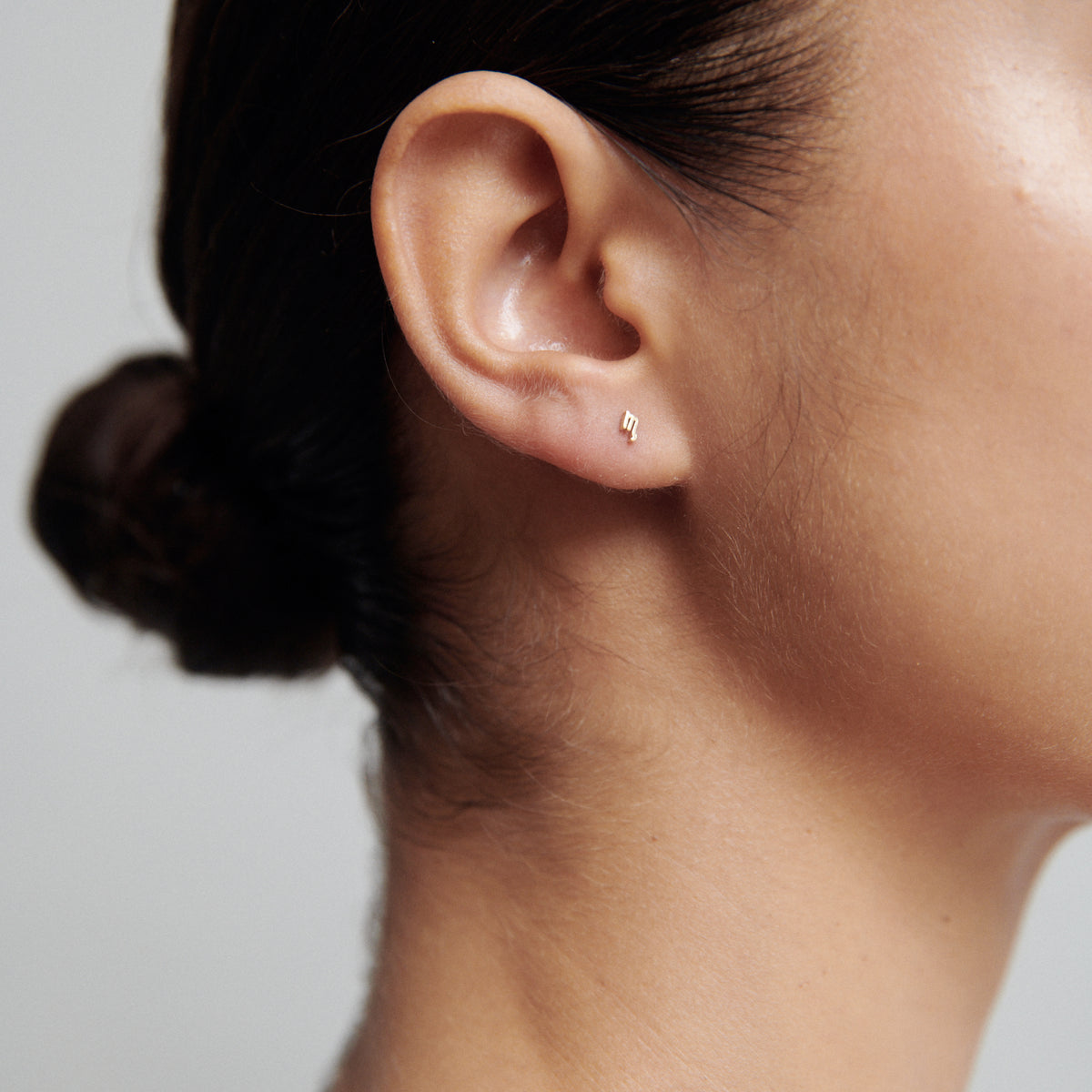 Close-up of a woman's ear wearing a small, elegant stud earring. Hair is tied in a bun, showcasing a minimalist jewellery style.