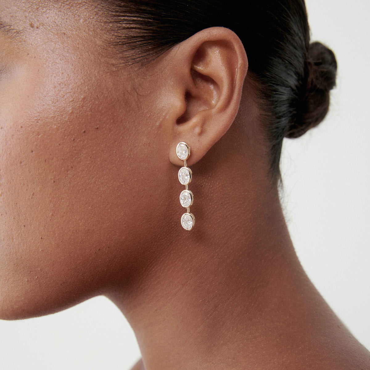Close-up of a woman's ear wearing elegant drop earrings with four oval diamonds, set in gold. Hair is tied back for a sophisticated look.