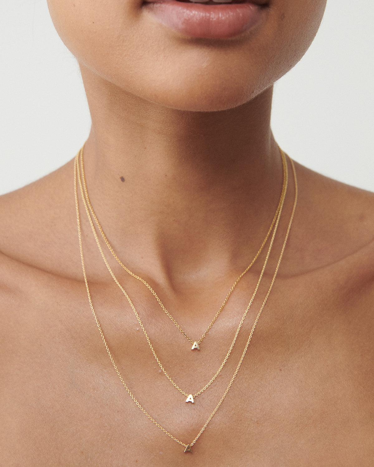 Close-up of a woman's neck wearing three delicate gold necklaces with small letter "A" pendants, layered elegantly for a minimalist look.