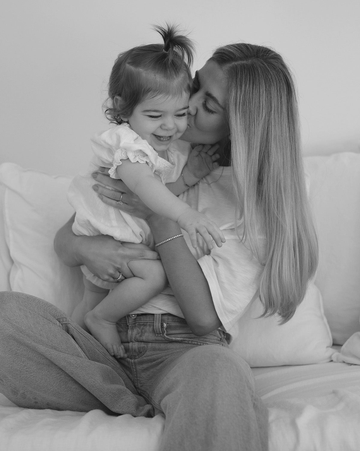 A mother lovingly kisses her smiling toddler, both in casual attire. The mother wears a delicate bracelet, highlighting luxury jewellery.