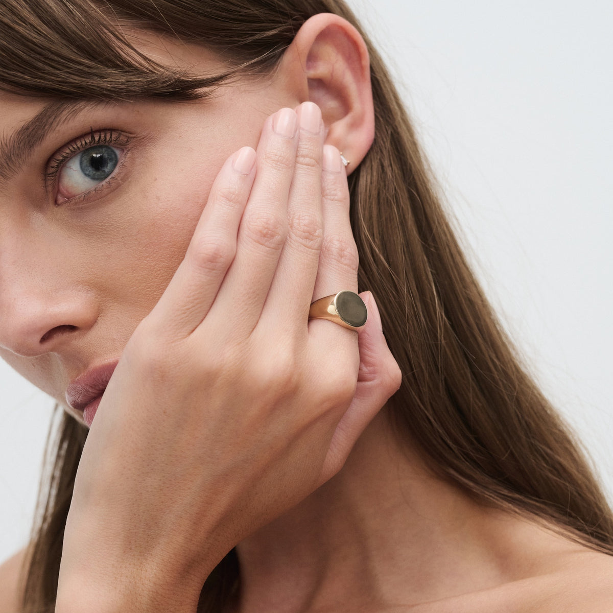 Woman with blue eyes wearing a gold signet ring, holding her hand to her face. Luxury jewellery focus, light background.