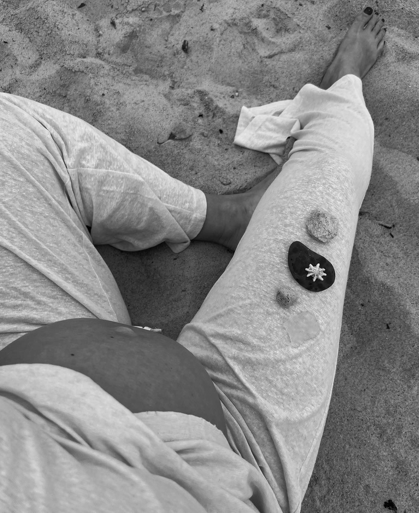 Person sitting on sand with stones and jewellery on their leg, wearing light clothing. Black and white photograph.