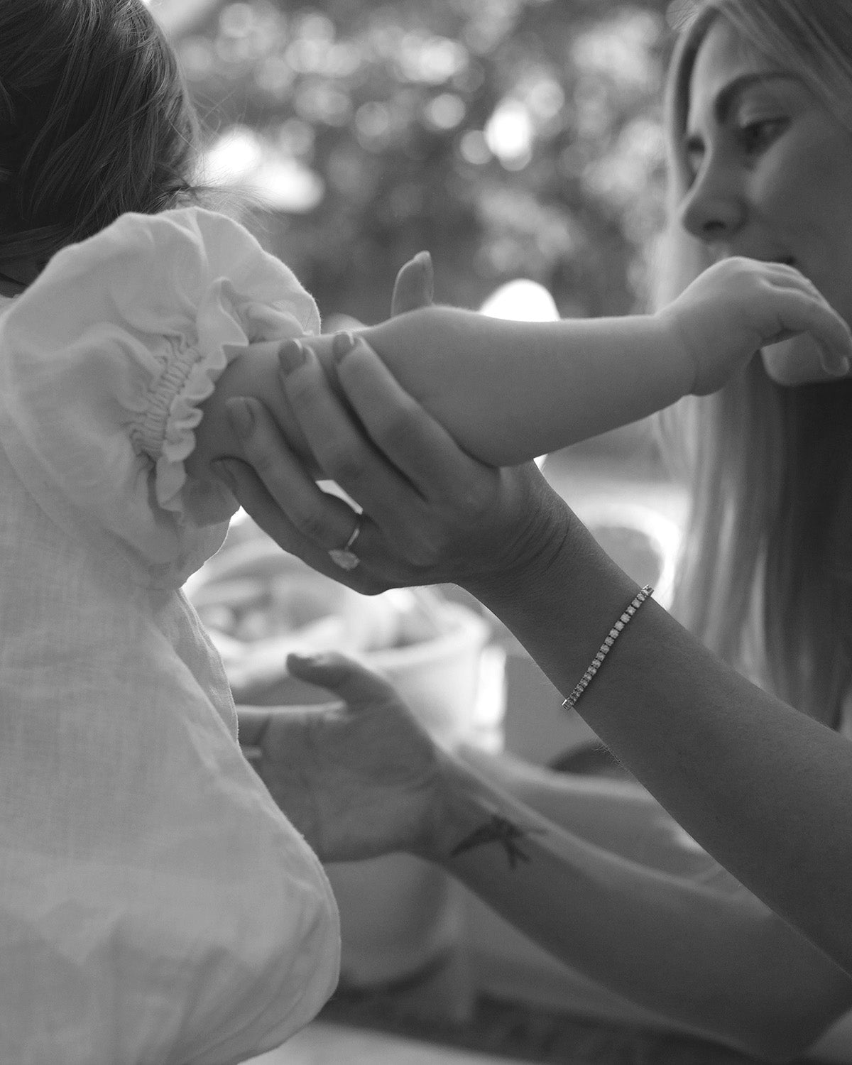 Woman in a white dress holds a baby’s arm, showcasing a delicate diamond bracelet. Soft focus and sunlight enhance the elegant scene.