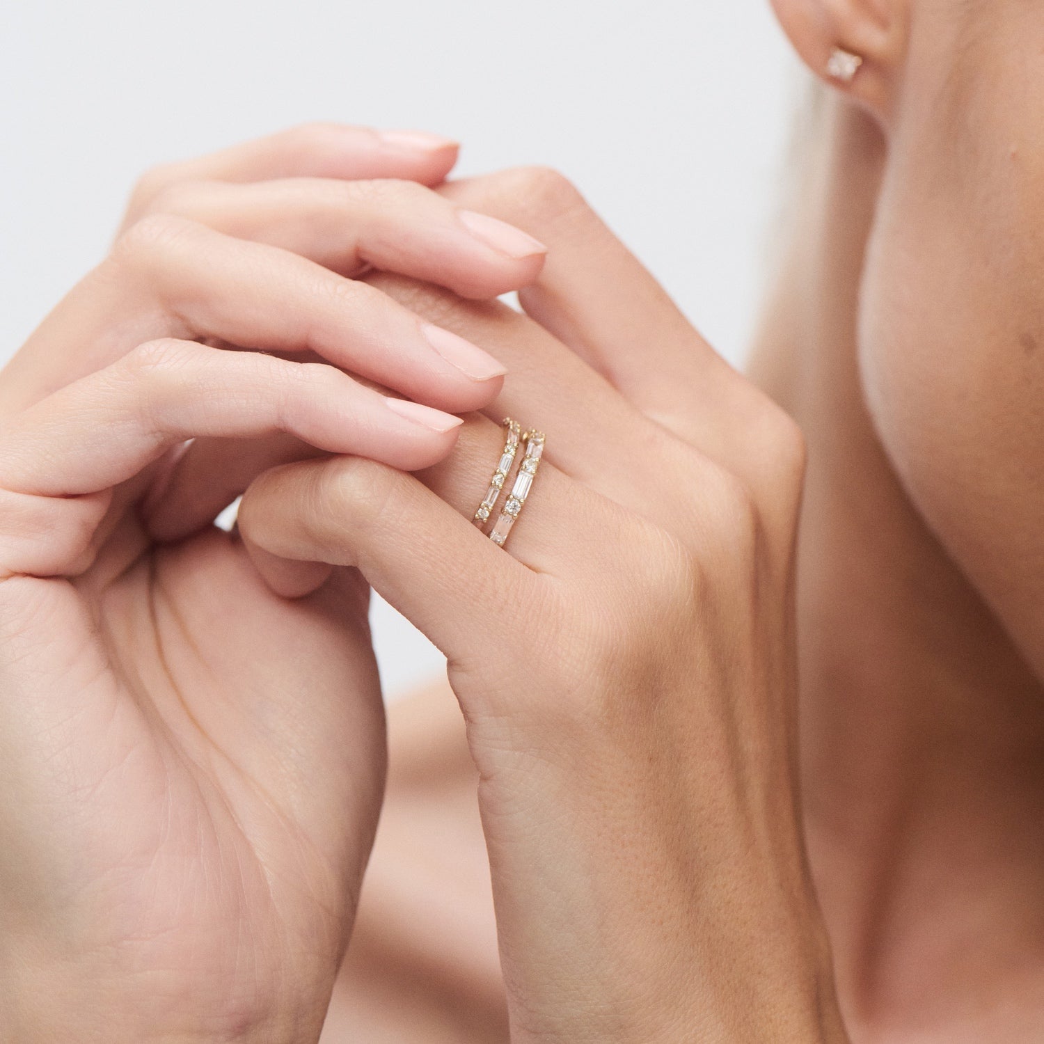 Elegant woman wearing stacked diamond rings on left hand, touching face. Luxury jewellery with gold bands and clear gemstones. Minimalist style. [text overlay:] 