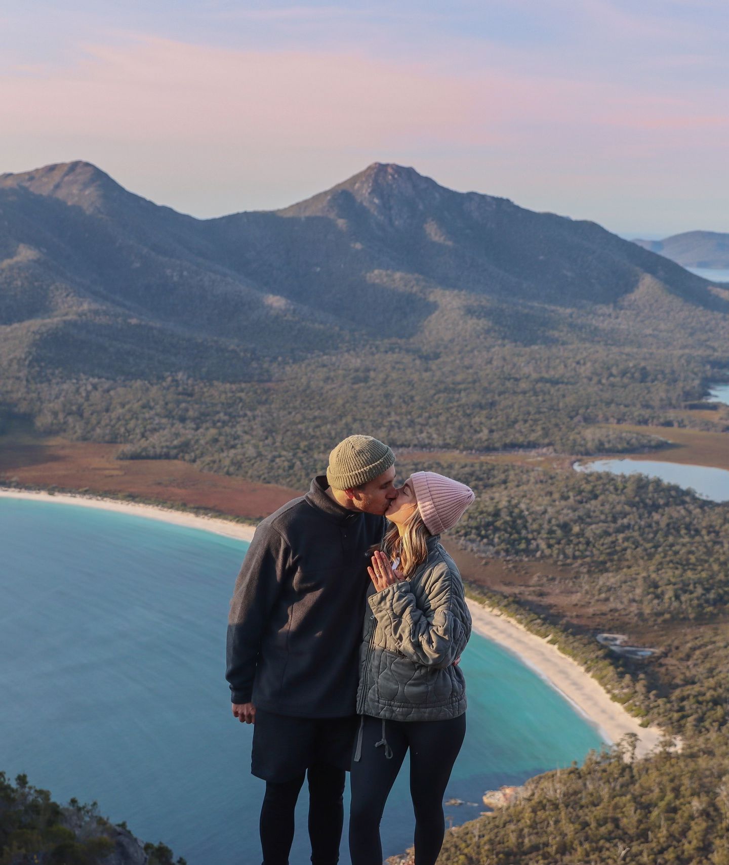 Couple kissing on a scenic mountaintop overlooking Wineglass Bay in Tasmania, with a beach and lush greenery below; a romantic, serene moment.
