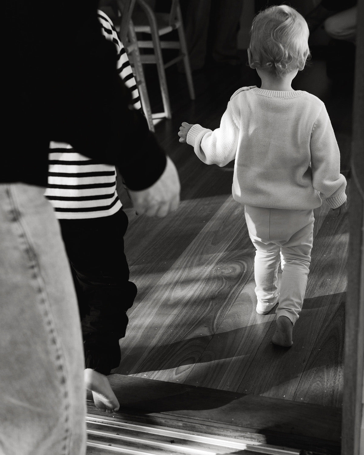 A child in a cosy sweater walks on wooden floorboards in a home, with adults nearby. Black and white photo showing a family moment.