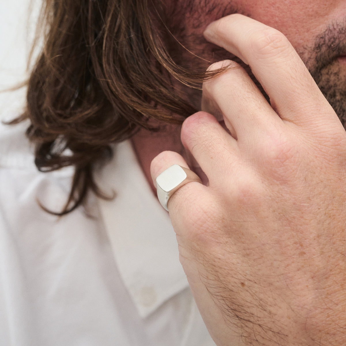 Man wearing a white shirt and silver signet ring, touching his face. Long brown hair visible. Luxury jewellery focus.