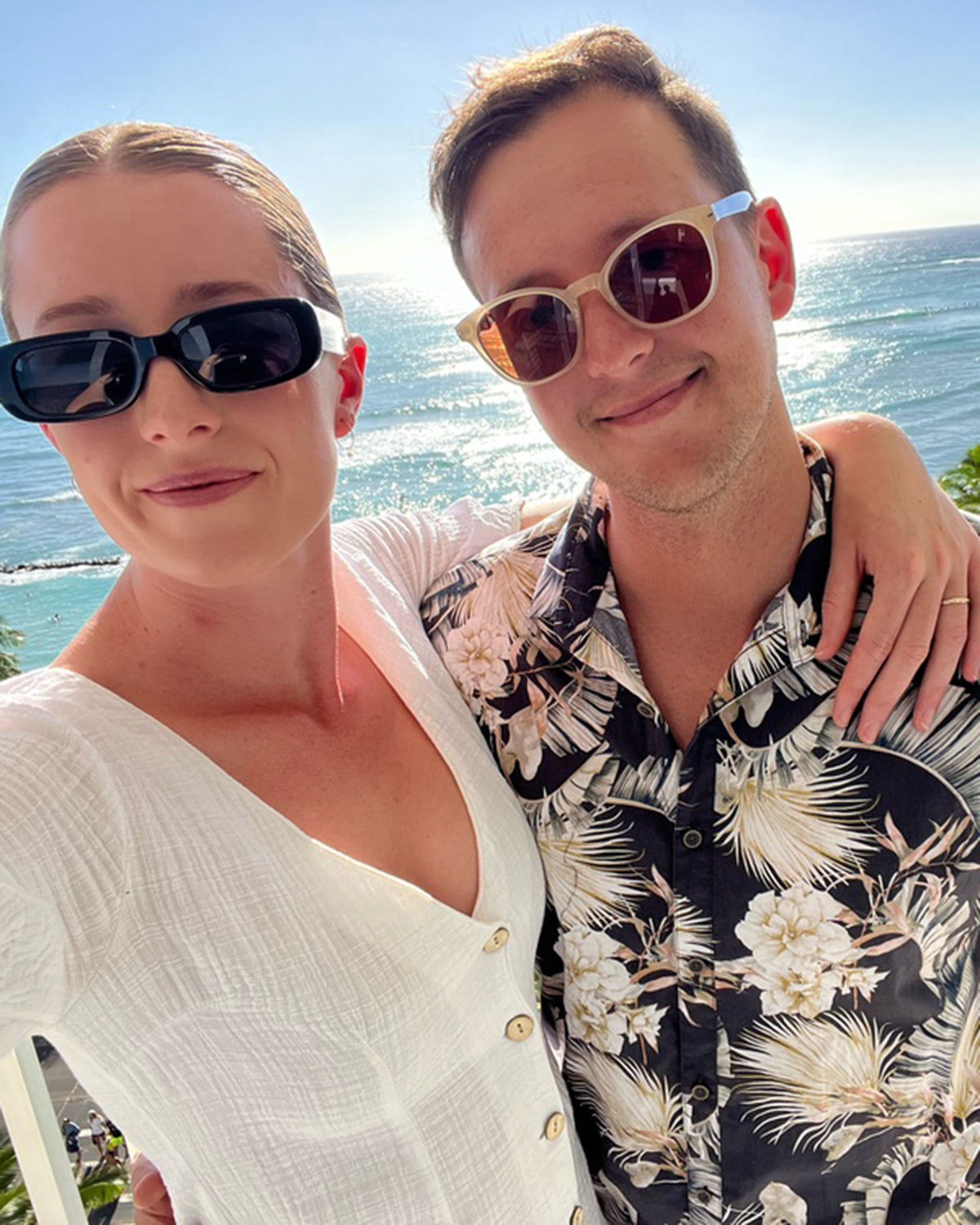 A couple in stylish sunglasses smiles at the camera, standing by the ocean, dressed in summery attire with a tropical backdrop.