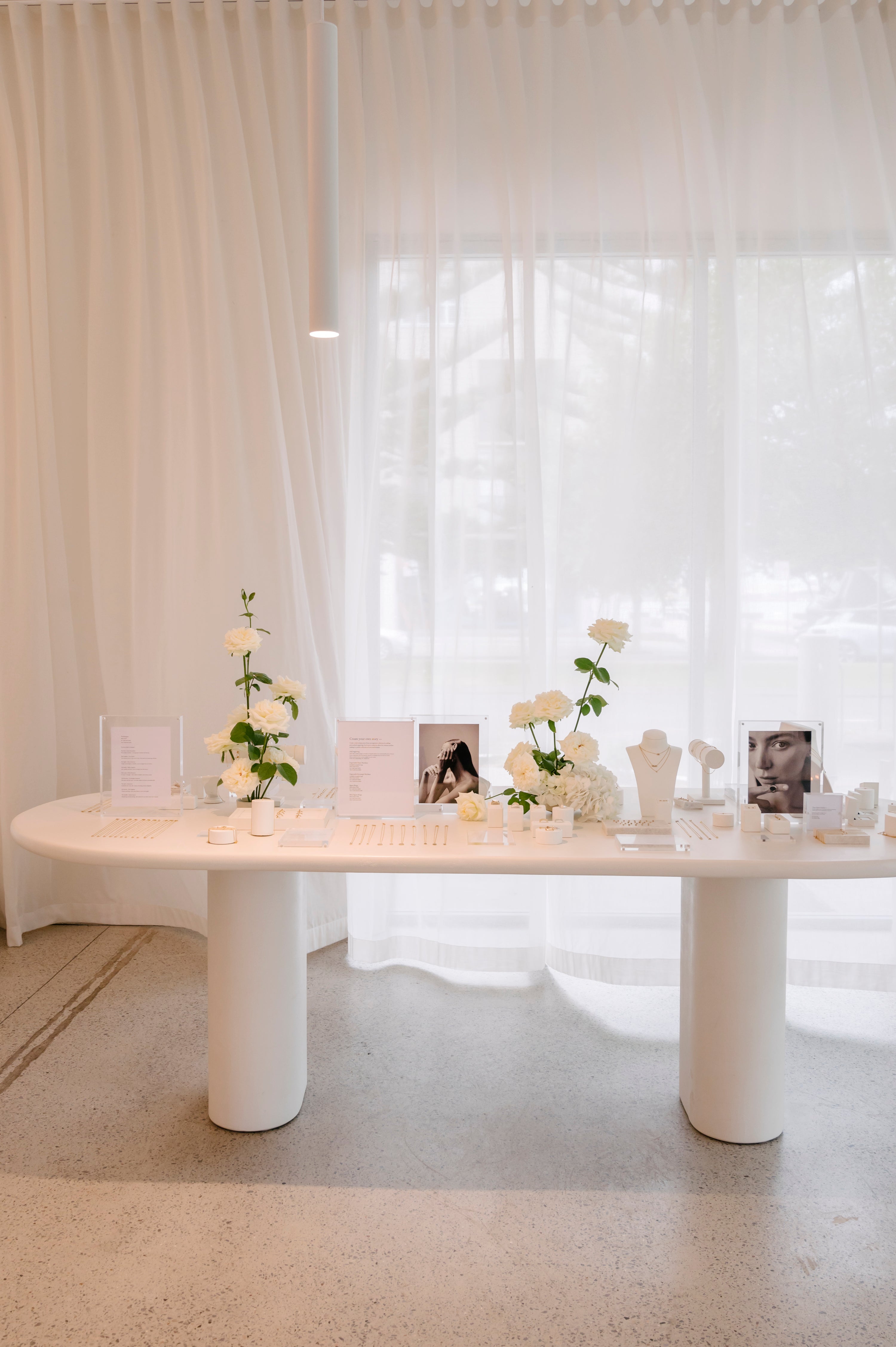 Elegant jewellery display on a white table with flowers, product photos, and white curtains in the background.
