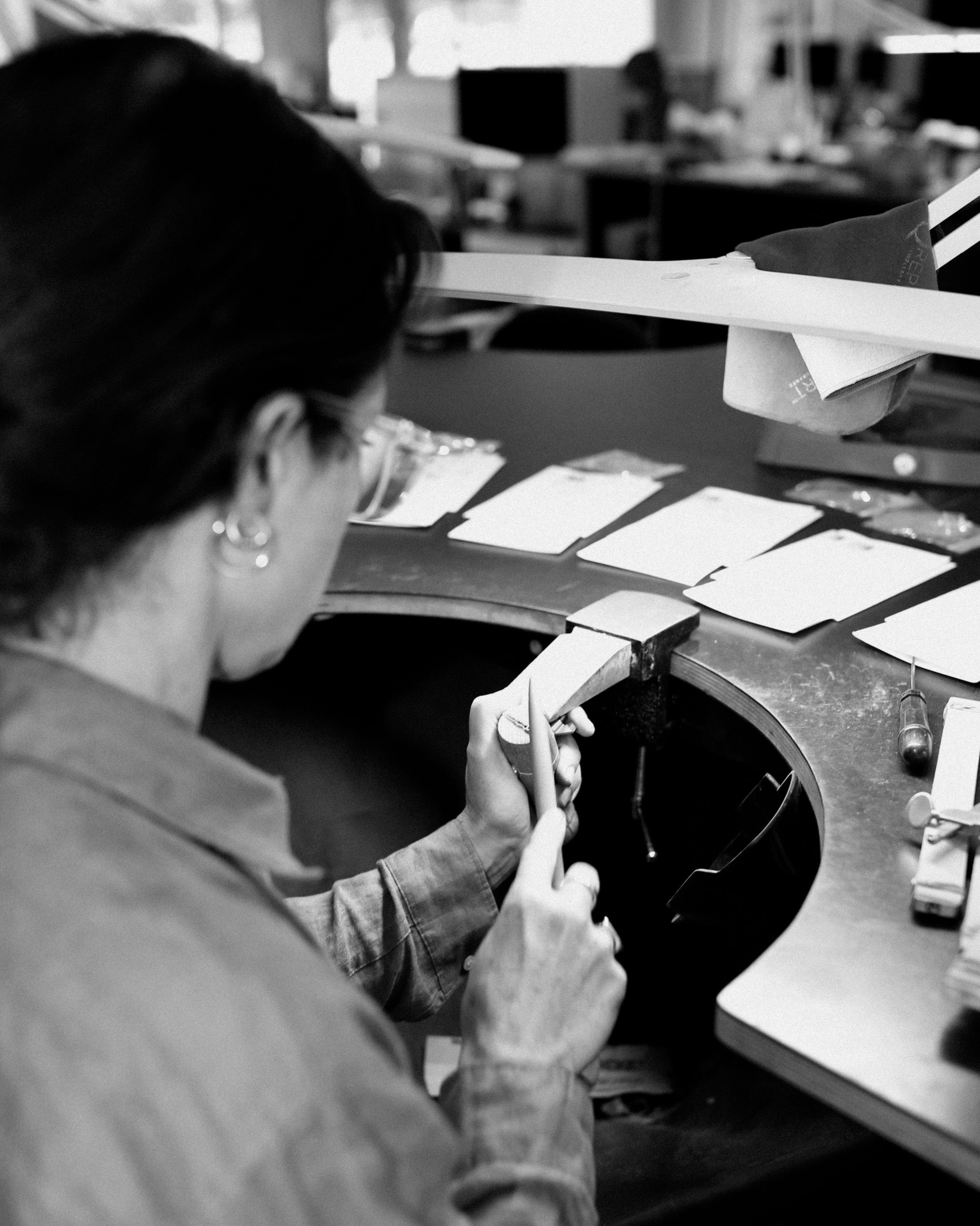 Jeweller crafting luxury pieces at a workbench, surrounded by tools and materials, in a high-end jewellery atelier. Monochrome image.