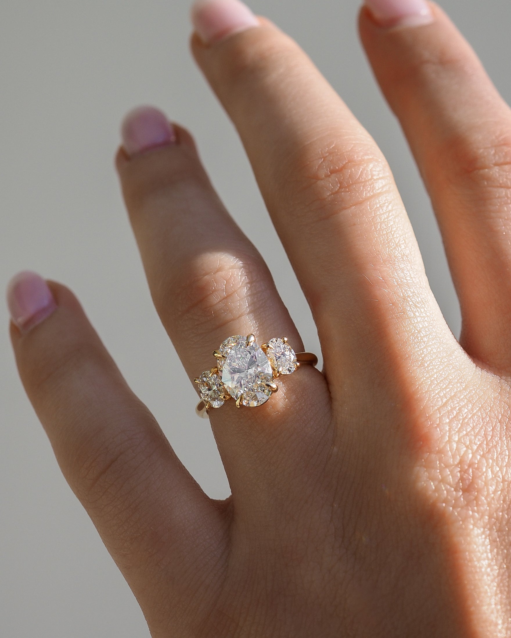 Gold ring with a large central oval diamond flanked by two smaller diamonds worn on a hand with light pink manicured nails.