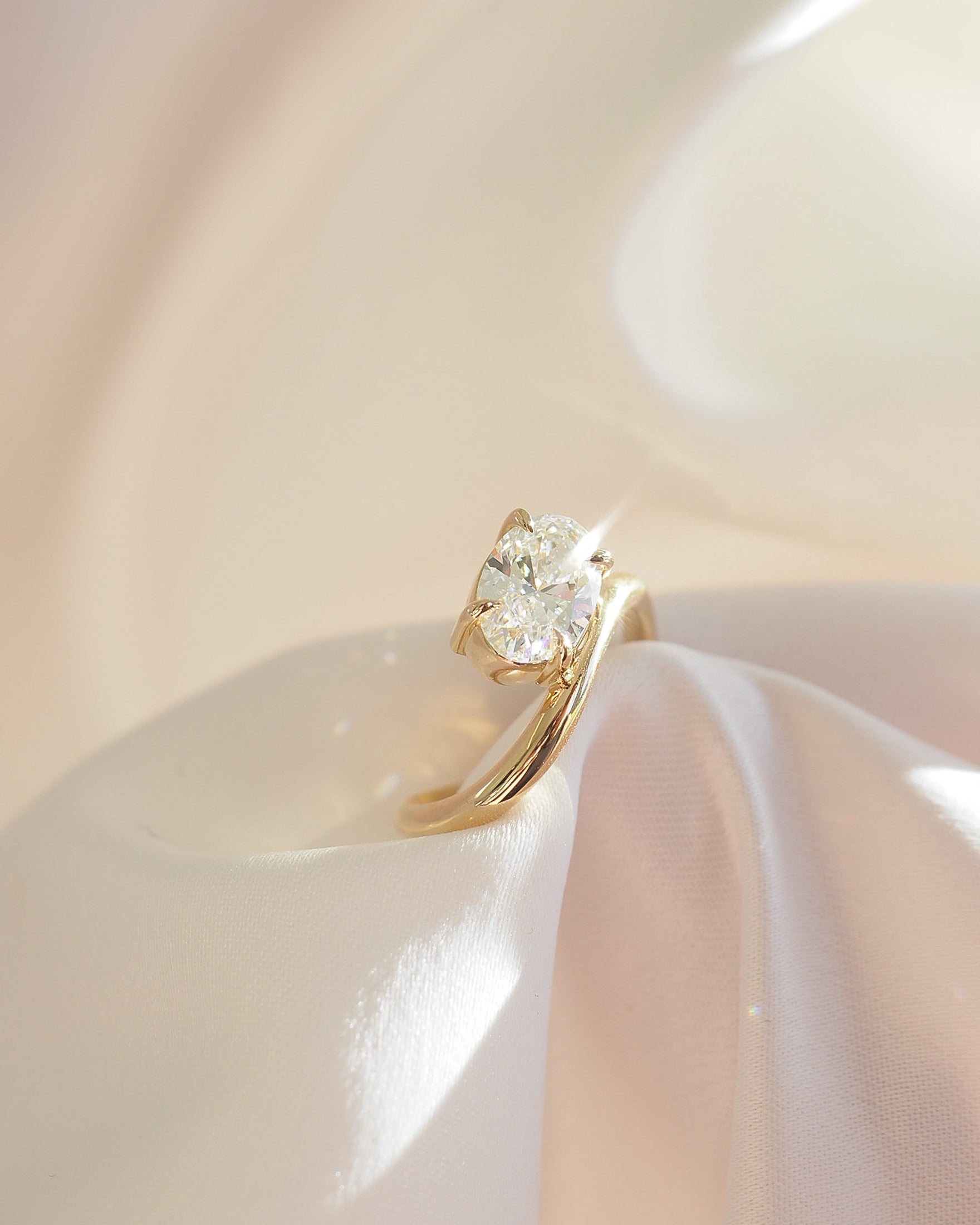 Elegant gold solitaire ring with a sparkling oval diamond, resting on soft, white fabric under gentle light. Luxurious jewellery spotlight.