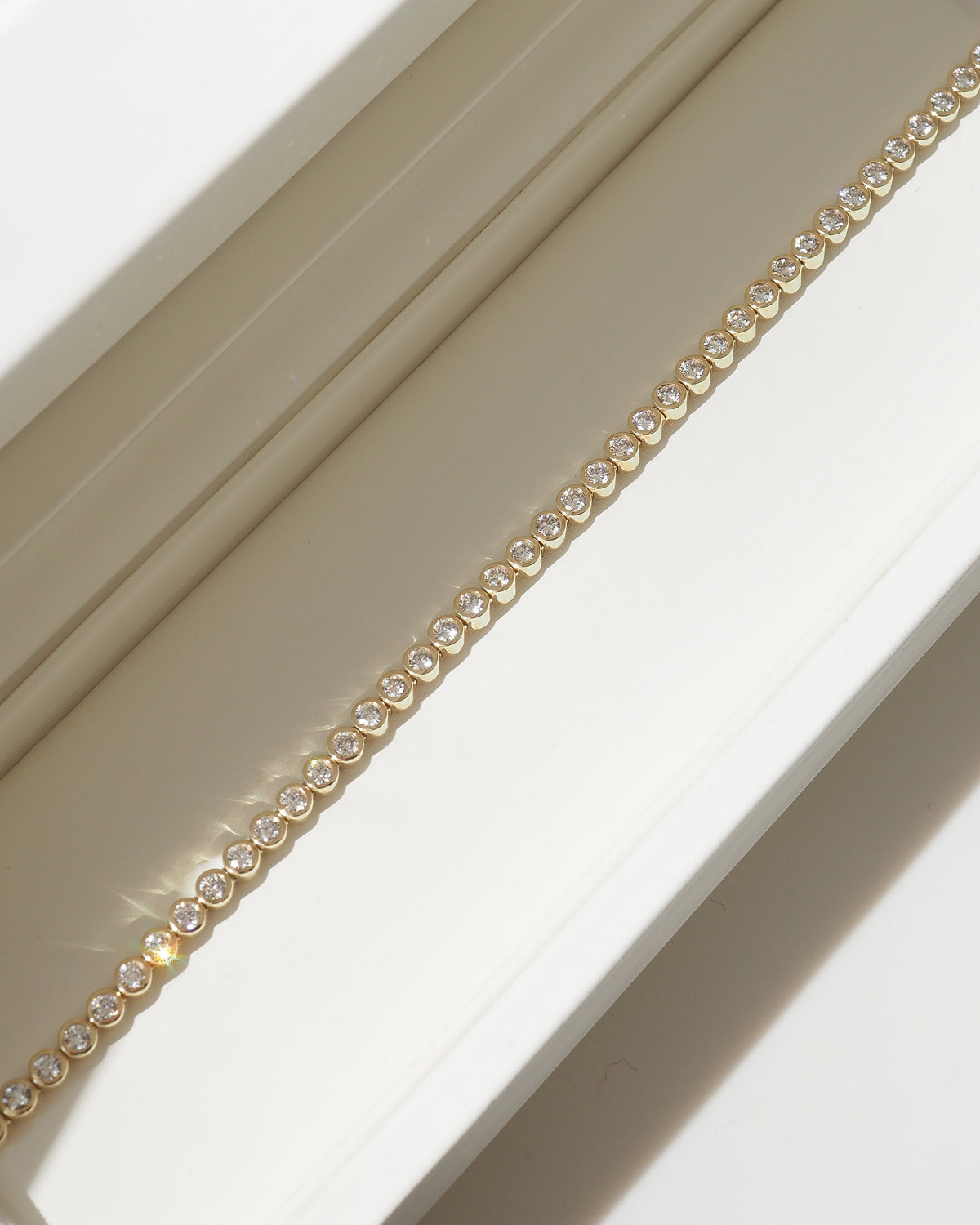 A close-up of a delicate gold chain bracelet with small clear round stones, laid diagonally across a cream-colored jewelry box. [text overlay: 2ct Bezel Tennis Bracelet] [text colour: #000000]