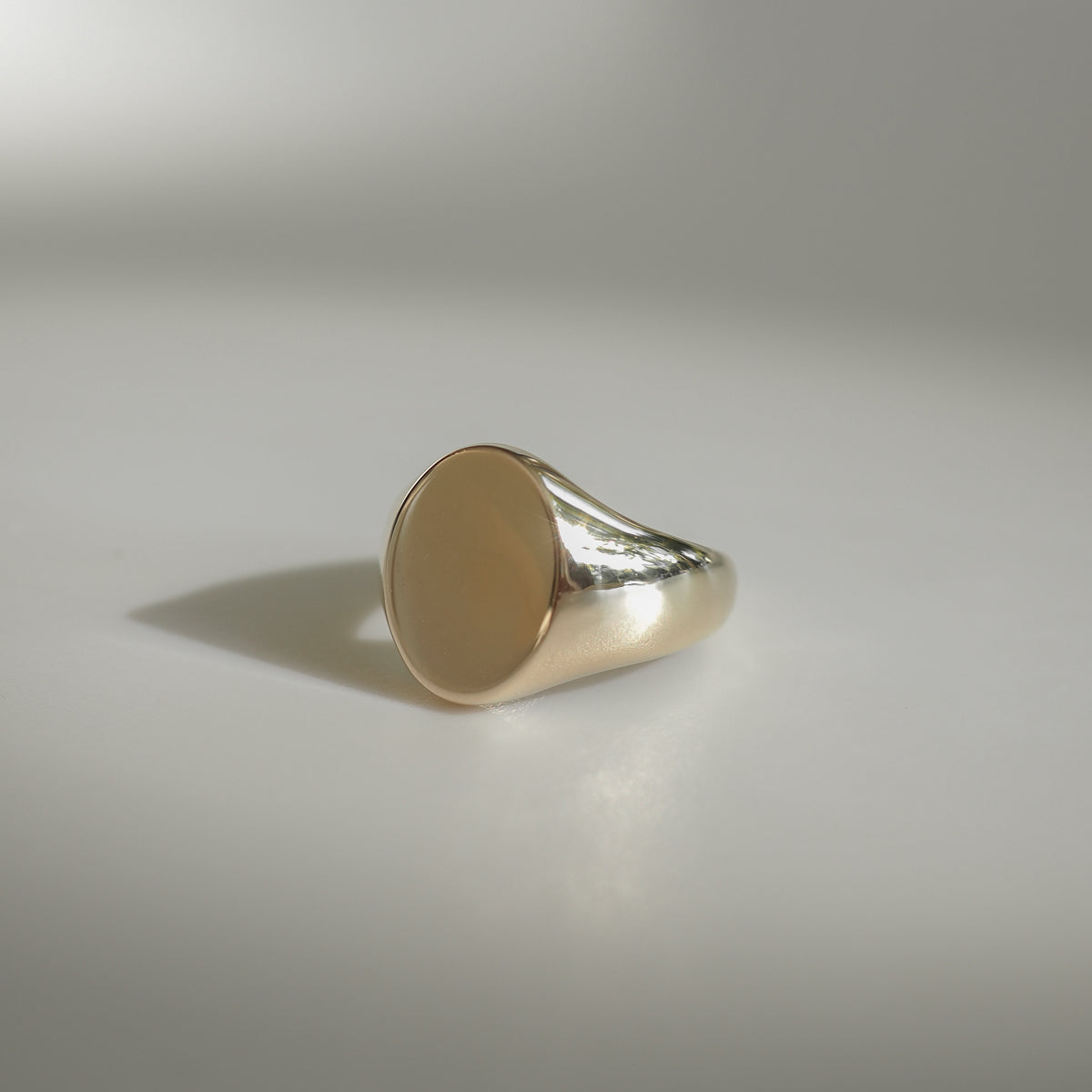 Gold signet ring with a smooth, polished oval face, reflecting light against a soft grey background. Elegant and classic design.