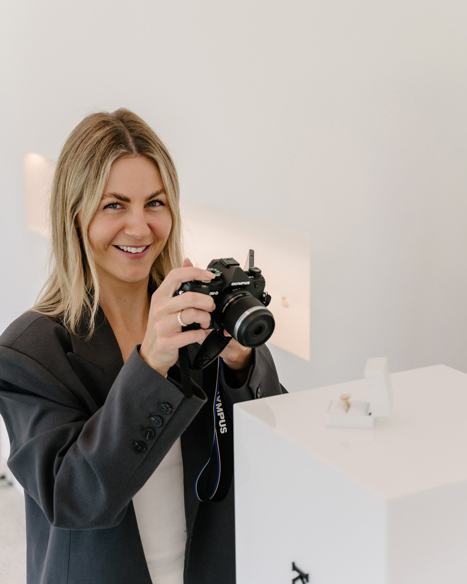 Woman photographing a luxury ring on display with a camera, dressed in a dark blazer, in a high-end jewellery store setting.