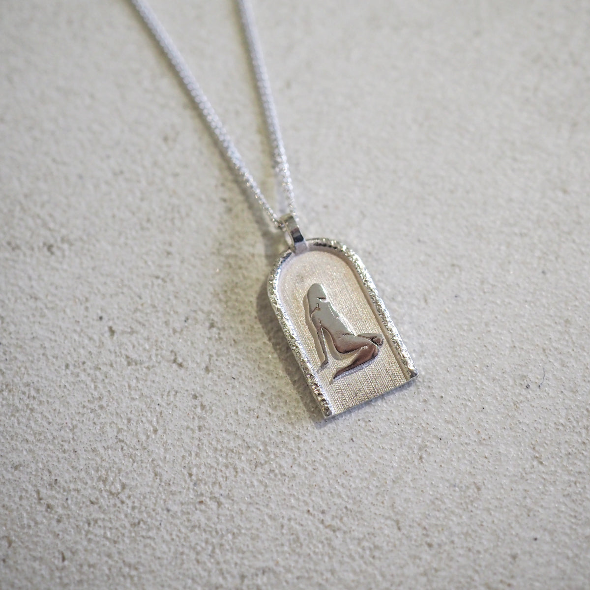 Silver pendant necklace featuring an engraved figure, on a textured beige surface. Luxurious high-end jewellery design.