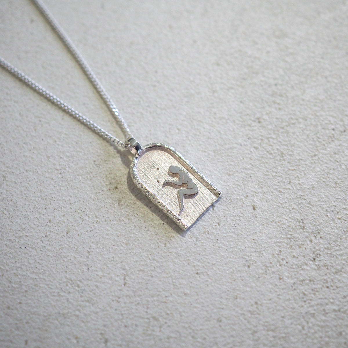 Silver pendant necklace with arch-shaped charm featuring a stylised figure design on textured surface, showcased on a light background.