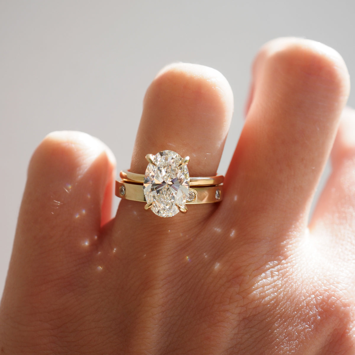 Hand with luxury gold ring and large oval diamond, paired with a matching gold band adorned with small diamonds. Sunlight adds sparkle.