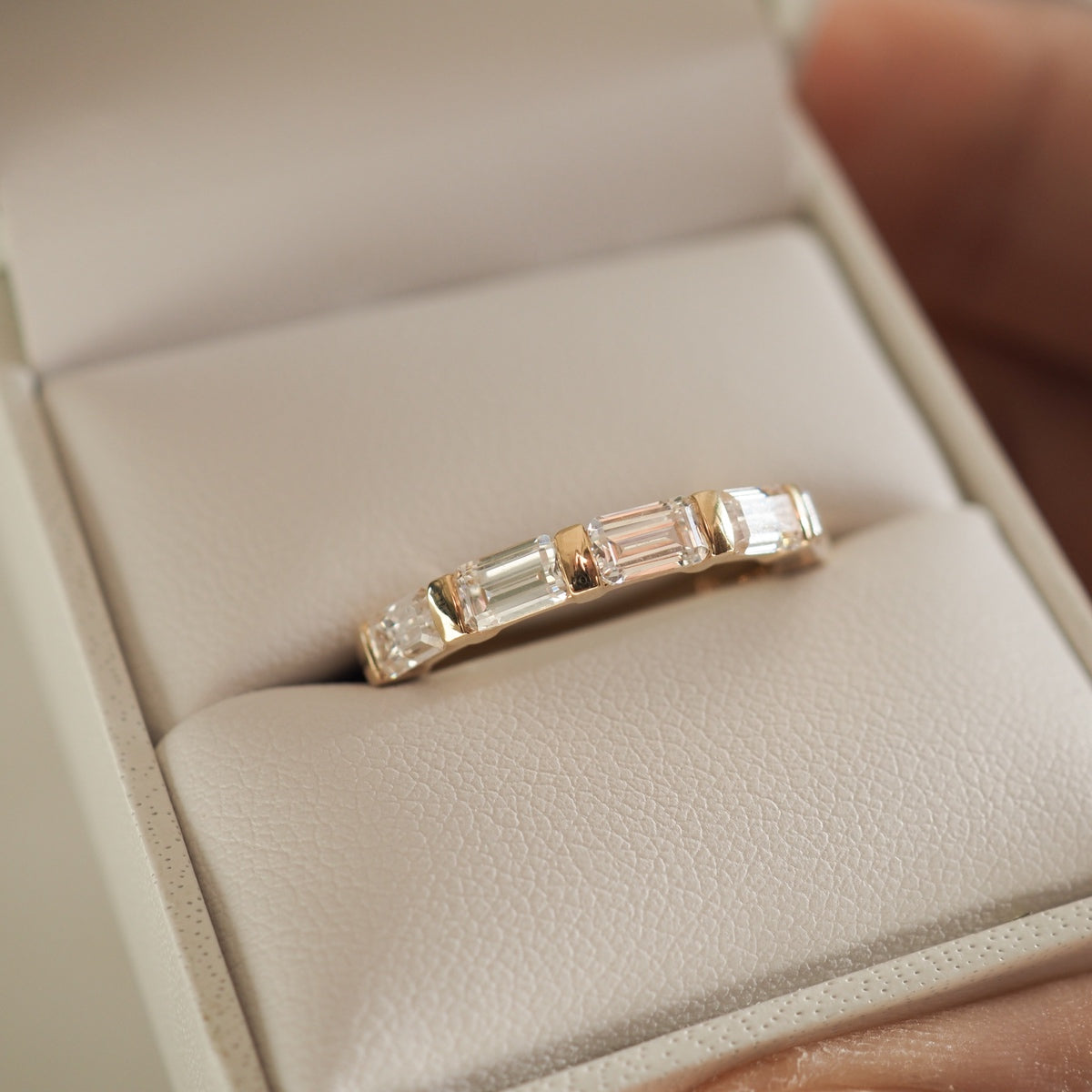 Gold ring with baguette-cut diamonds, displayed in a beige jewellery box. Elegant and luxurious design. Ideal for weddings or special occasions.