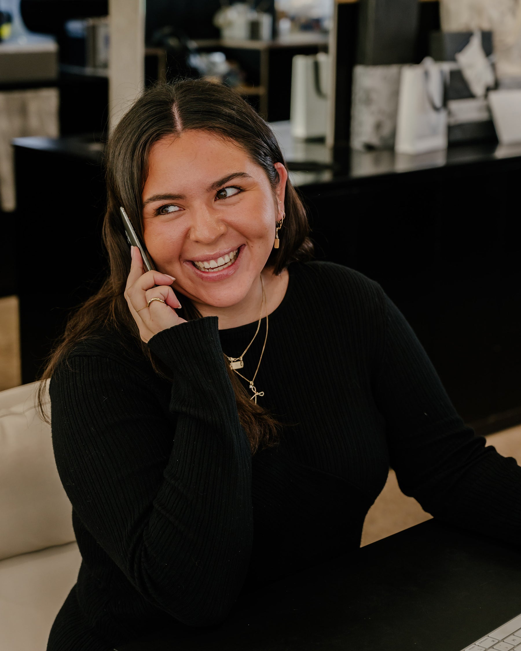 Smiling woman on phone in luxury jewellery store, wearing black sweater and gold jewellery, seated at a black counter.