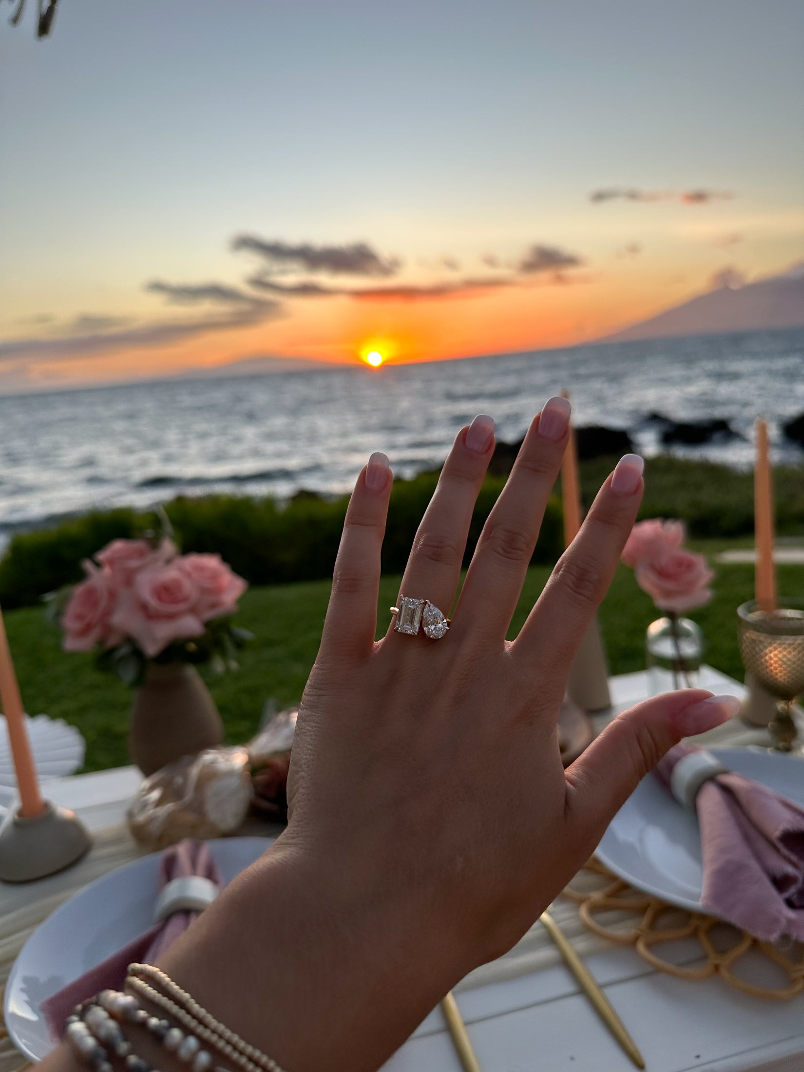 Hand wearing elegant diamond ring with luxury wedding band, set against a romantic beach sunset. Roses and candles adorn a lavish dining table.