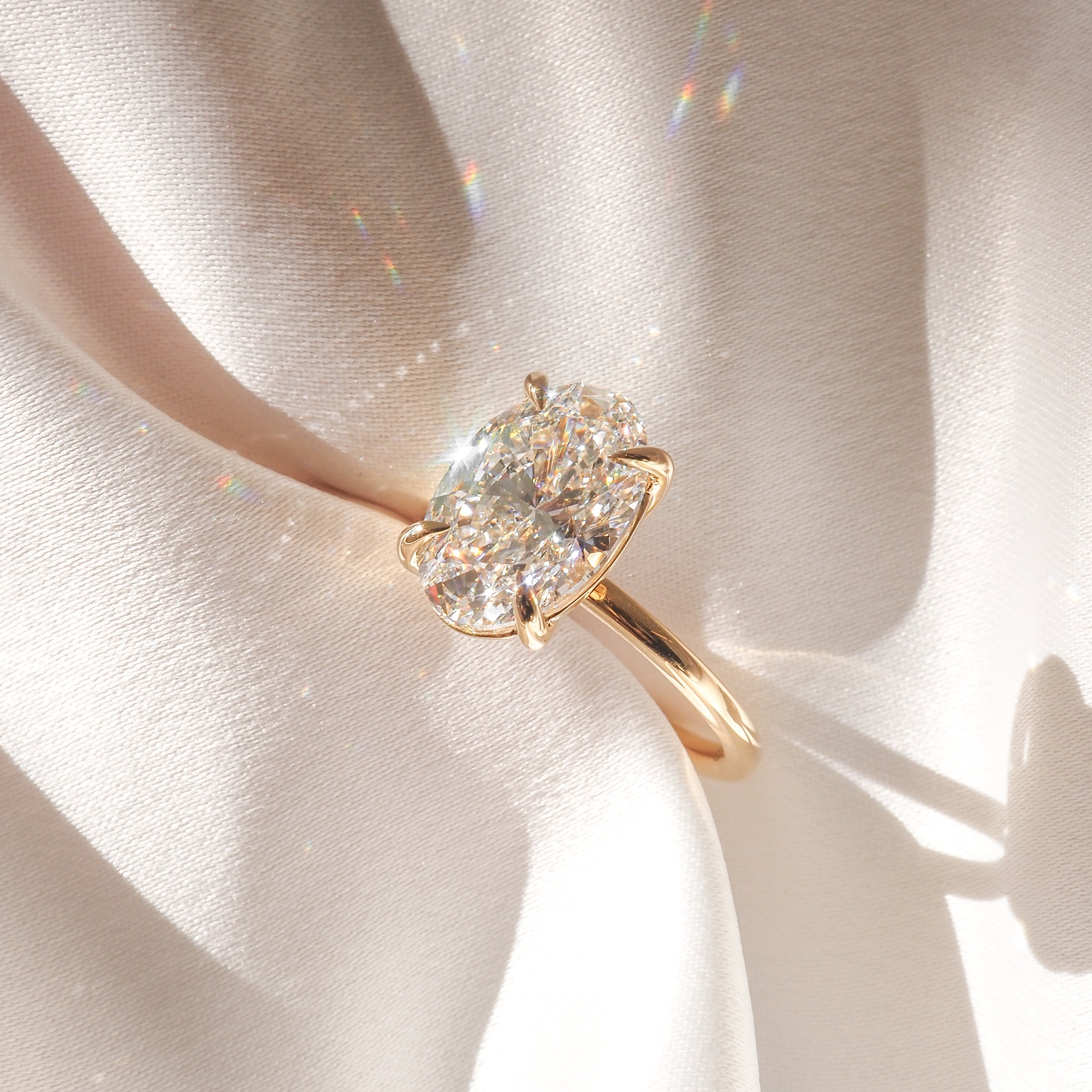 Elegant gold ring with a large oval diamond set against luxurious beige silk, reflecting rainbow light.