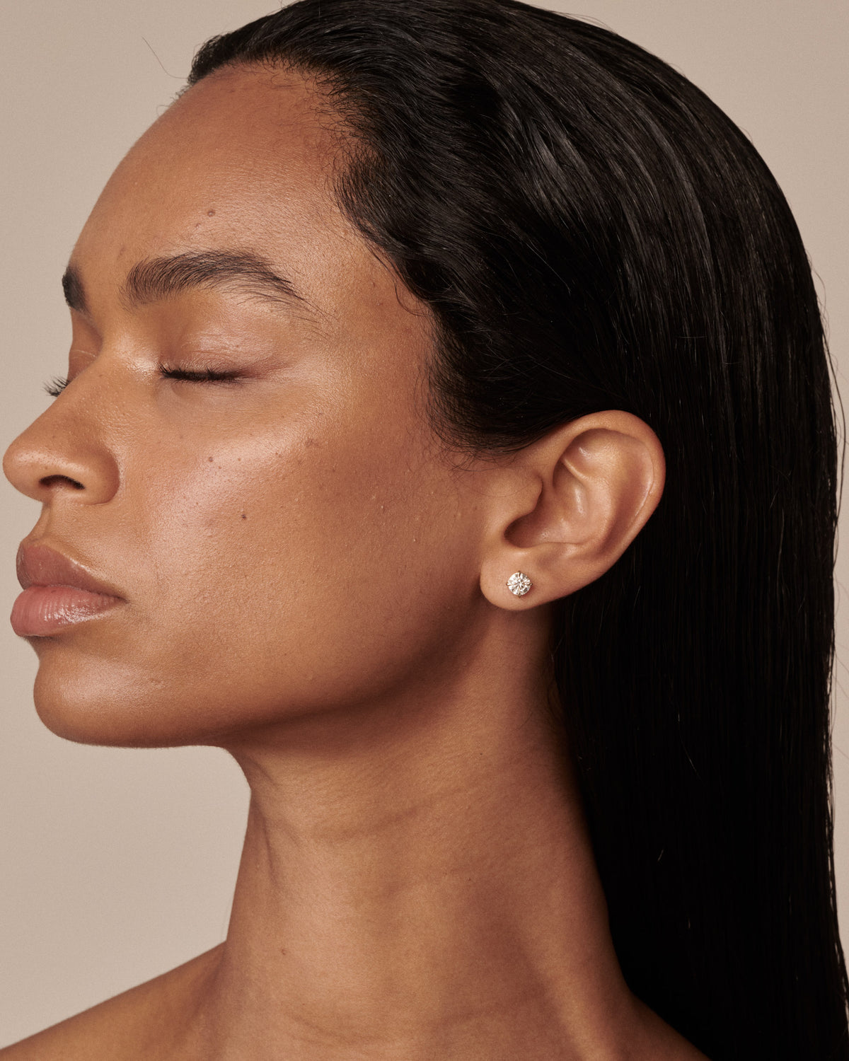 A close-up side profile of a woman with closed eyes wearing a small round silver stud earring with a clear diamond, set against a solid beige background.