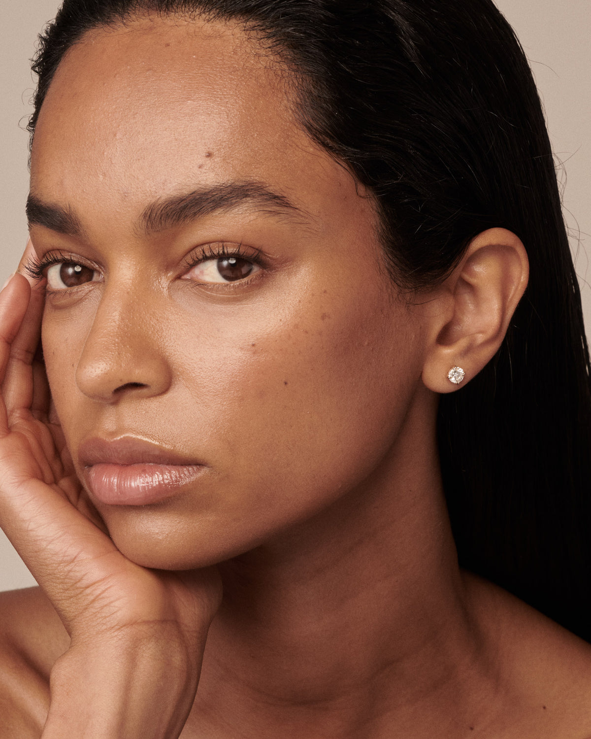 A close-up portrait of a woman with dark hair, wearing a small round silver stud earring with a clear diamond, set against a warm beige background. [text overlay: The Cleo Studs in 1.5ct (0.75ct each)]