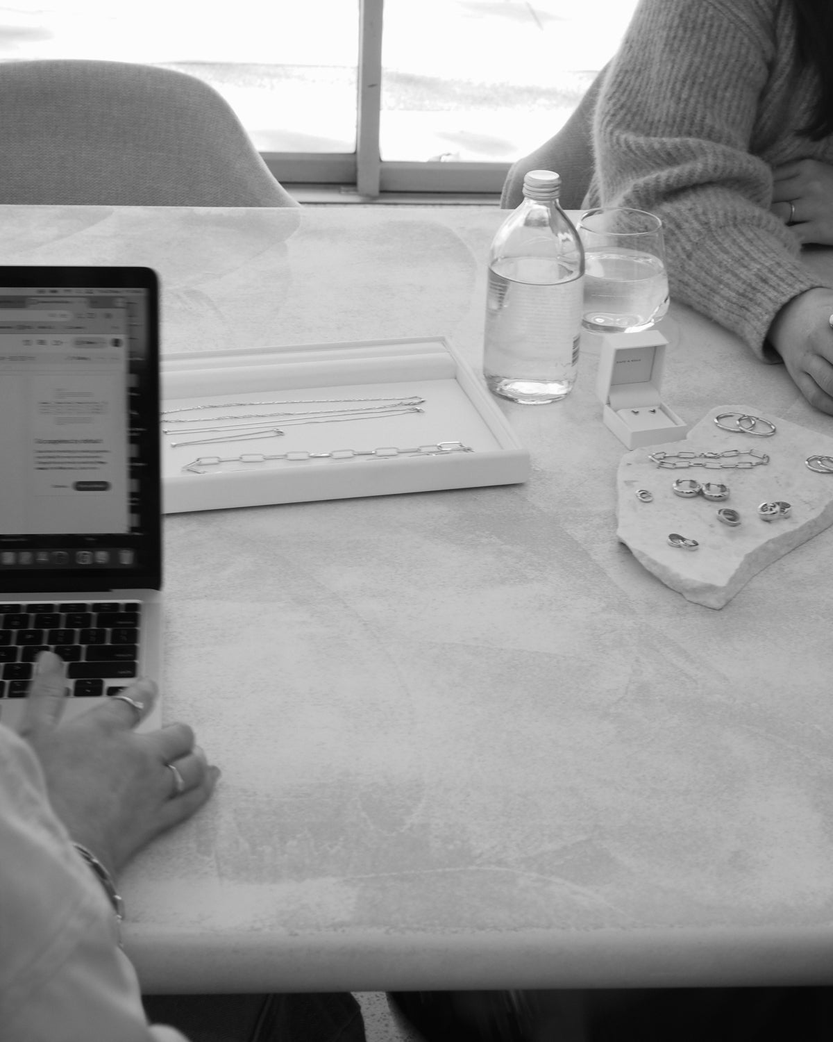 A marble table displays luxury jewellery with a laptop, water bottle, and wine glass. People are partially visible, suggesting a meeting or review.