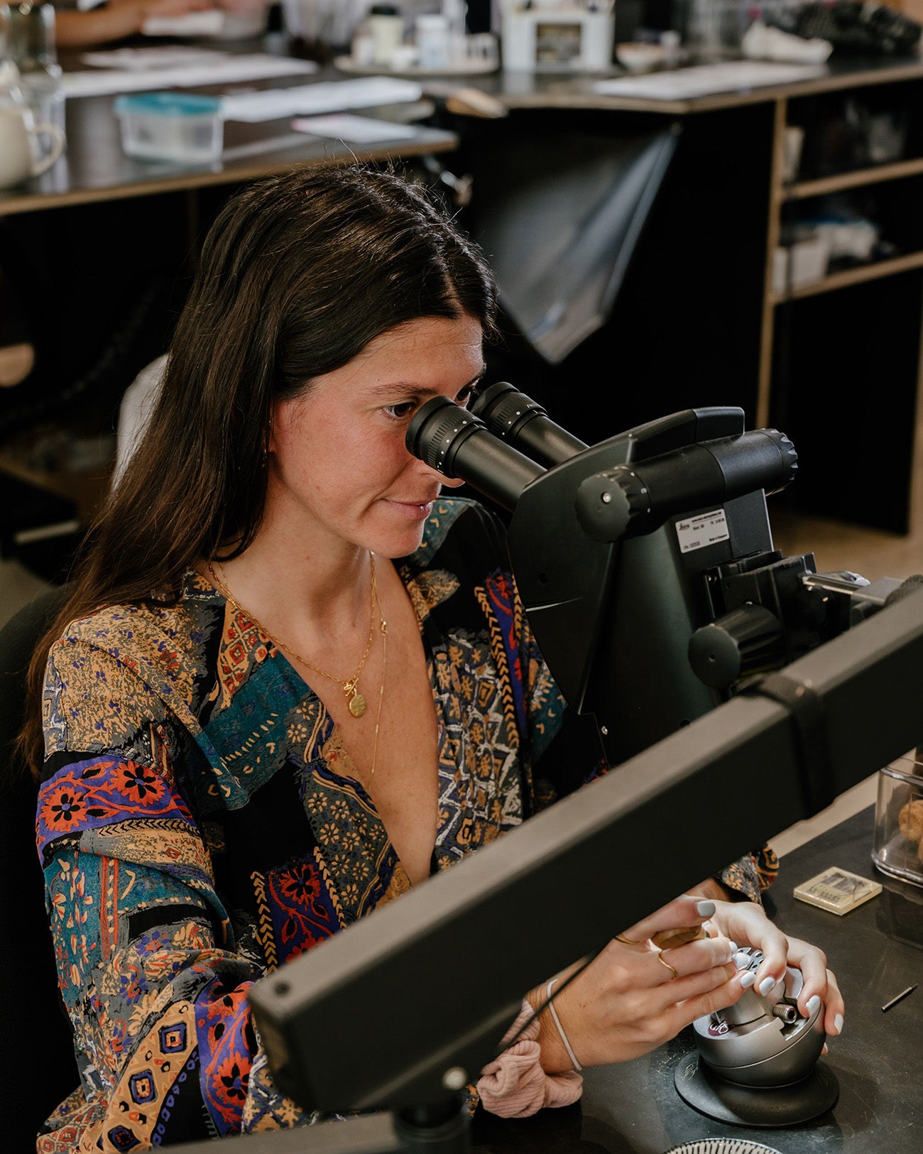 Woman examining jewellery through a microscope in a workshop, wearing a colourful patterned blouse. Luxurious jewellery craftsmanship scene.
