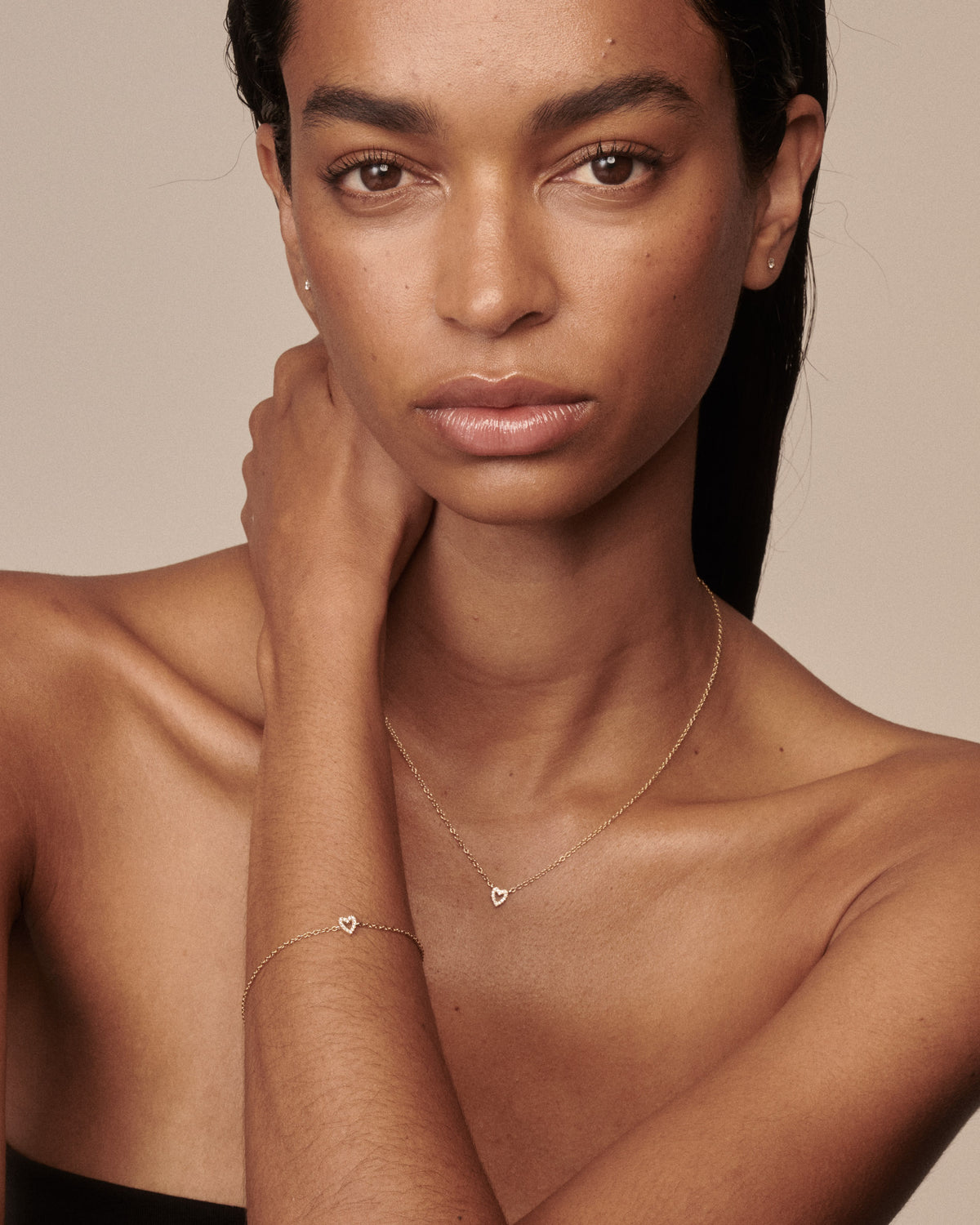 A close-up portrait of a woman wearing delicate gold jewelry, including a thin chain necklace with a small heart pendant and a matching bracelet with a small heart charm, against a warm beige background.