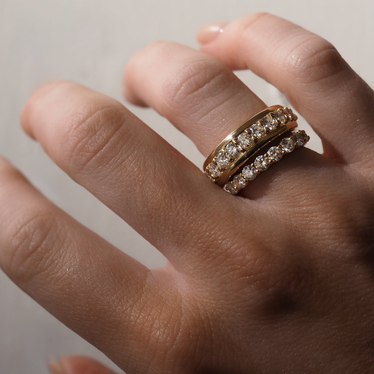 Two gold rings with sparkling diamonds adorning a person's fingers against a neutral background, showcasing luxury jewellery style.