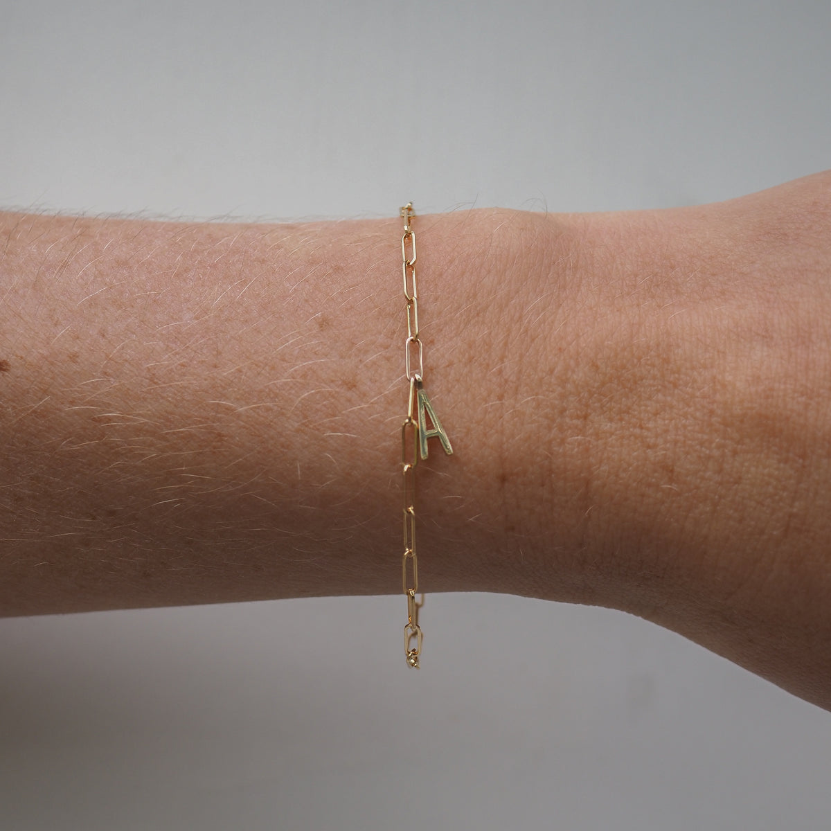 Gold chain bracelet with an 'A' initial pendant on a wrist, against a plain background. Minimalist, luxury jewellery design.