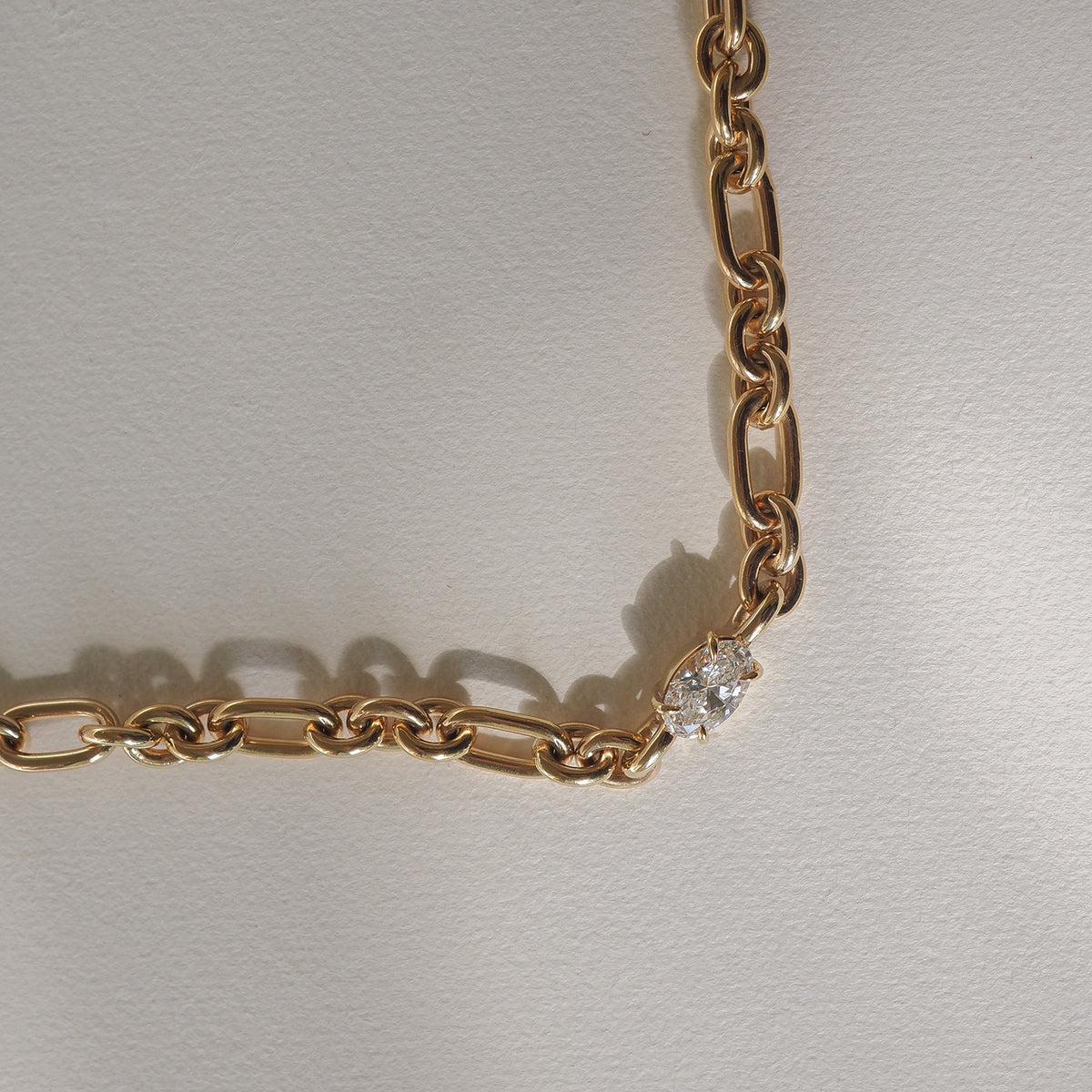 Gold chain necklace with a single sparkling diamond link, casting soft shadows on a cream surface. Luxurious jewellery design.