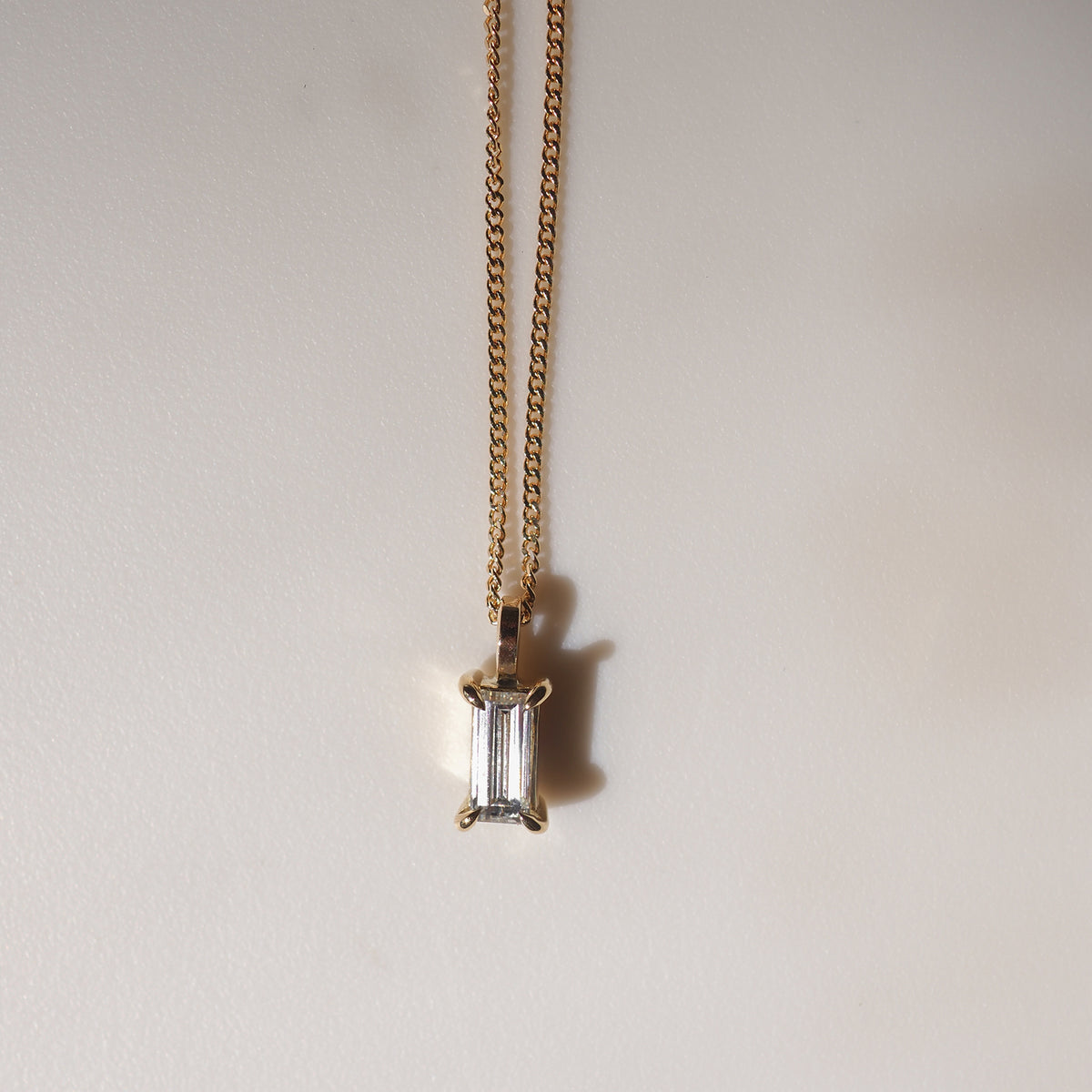 Gold chain necklace with an emerald-cut diamond pendant against a plain background, casting a subtle shadow. Luxurious and elegant design.