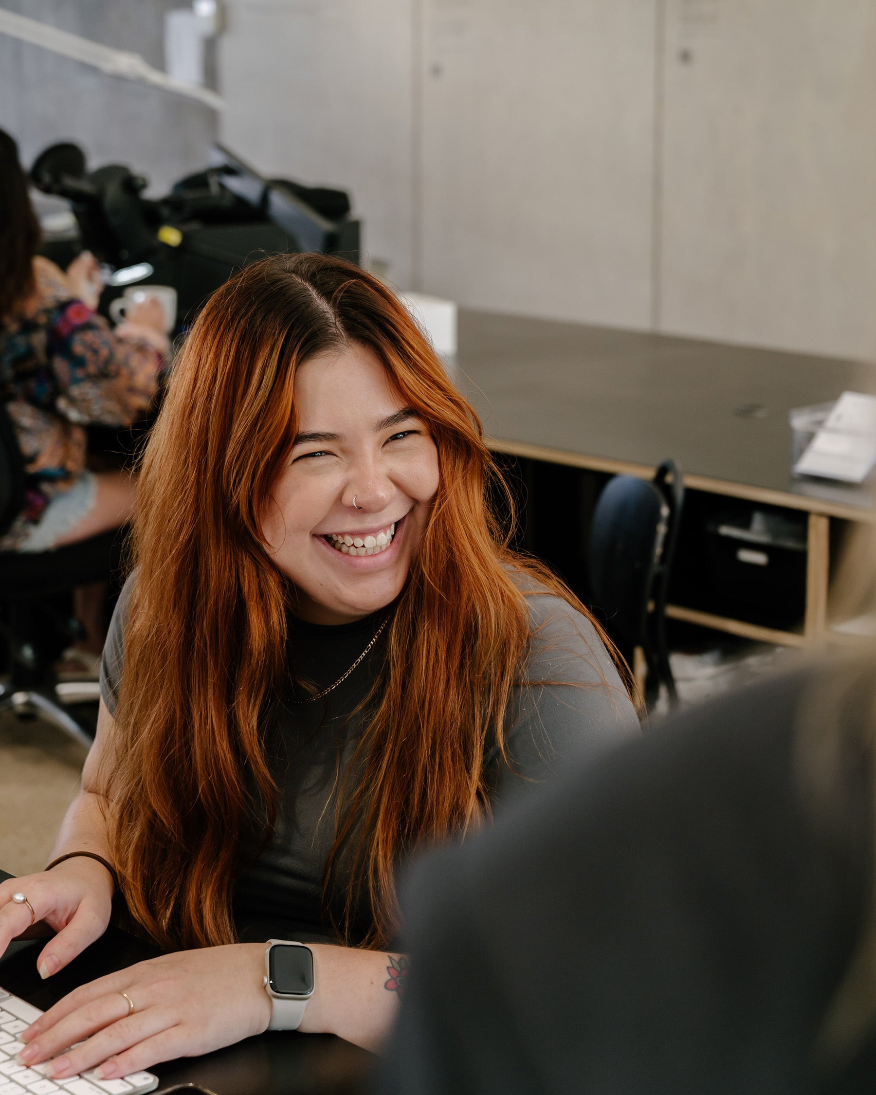 Smiling woman with long red hair at a desk, wearing a smartwatch. Casual office setting, blurred background; modern workplace vibe.