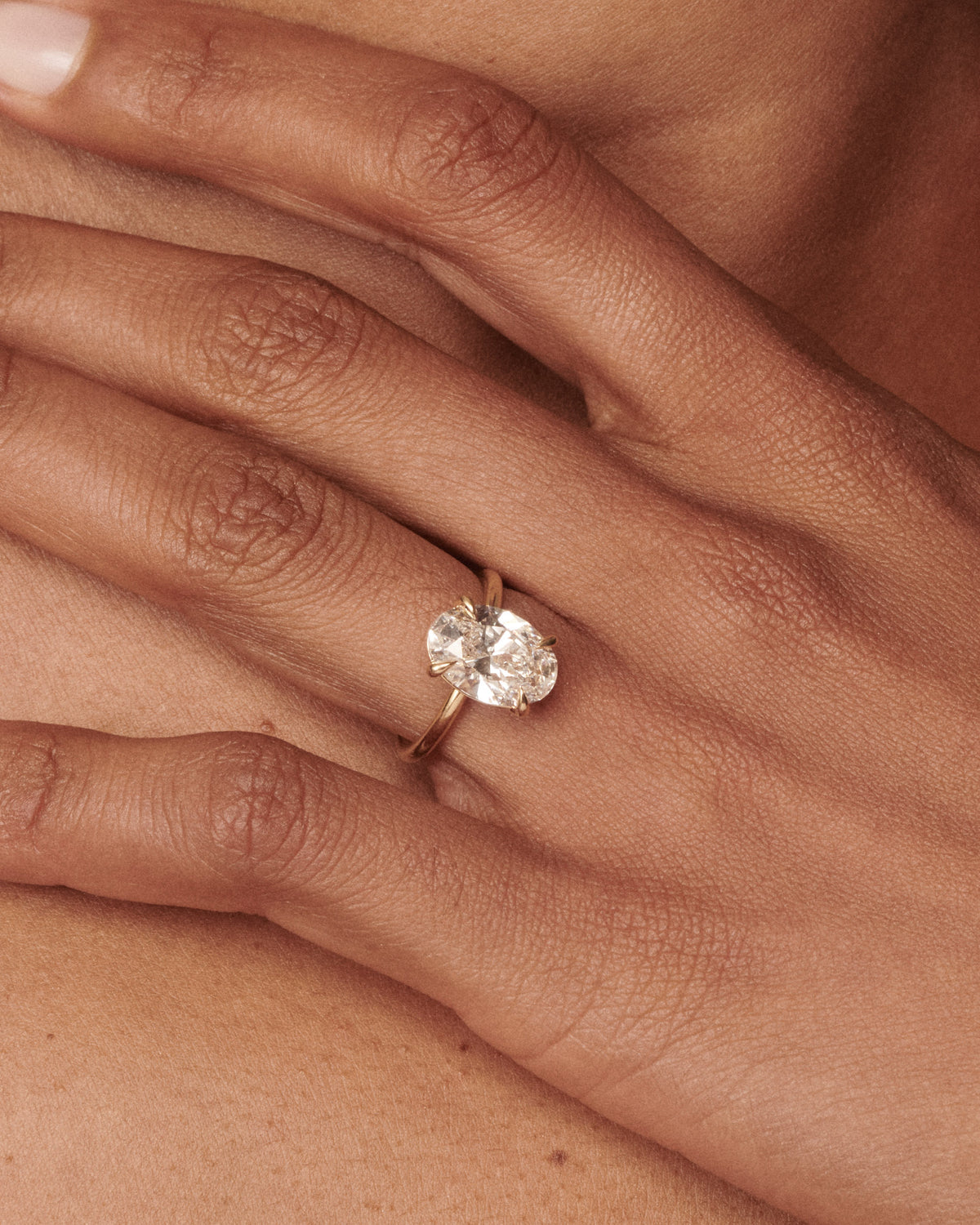 A close-up of a person's hand wearing a yellow gold ring with an oval-cut clear gemstone, set against a soft beige background.