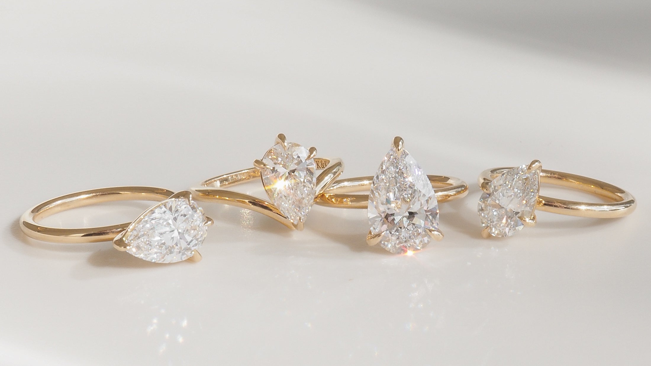 Four gold rings with pear-shaped diamonds elegantly displayed on a smooth, reflective surface, showcasing luxury jewellery design.
