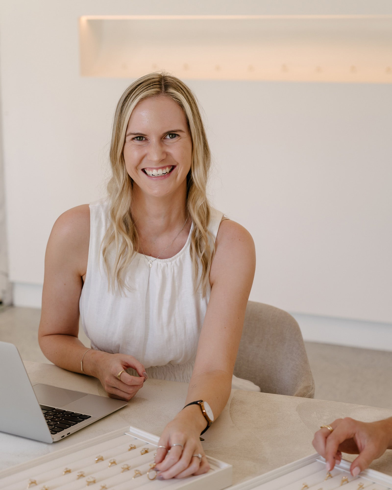 Woman in white dress smiling, selecting rings from a jewellery tray, sitting at a table with a laptop. Elegant, modern jewellery setting.