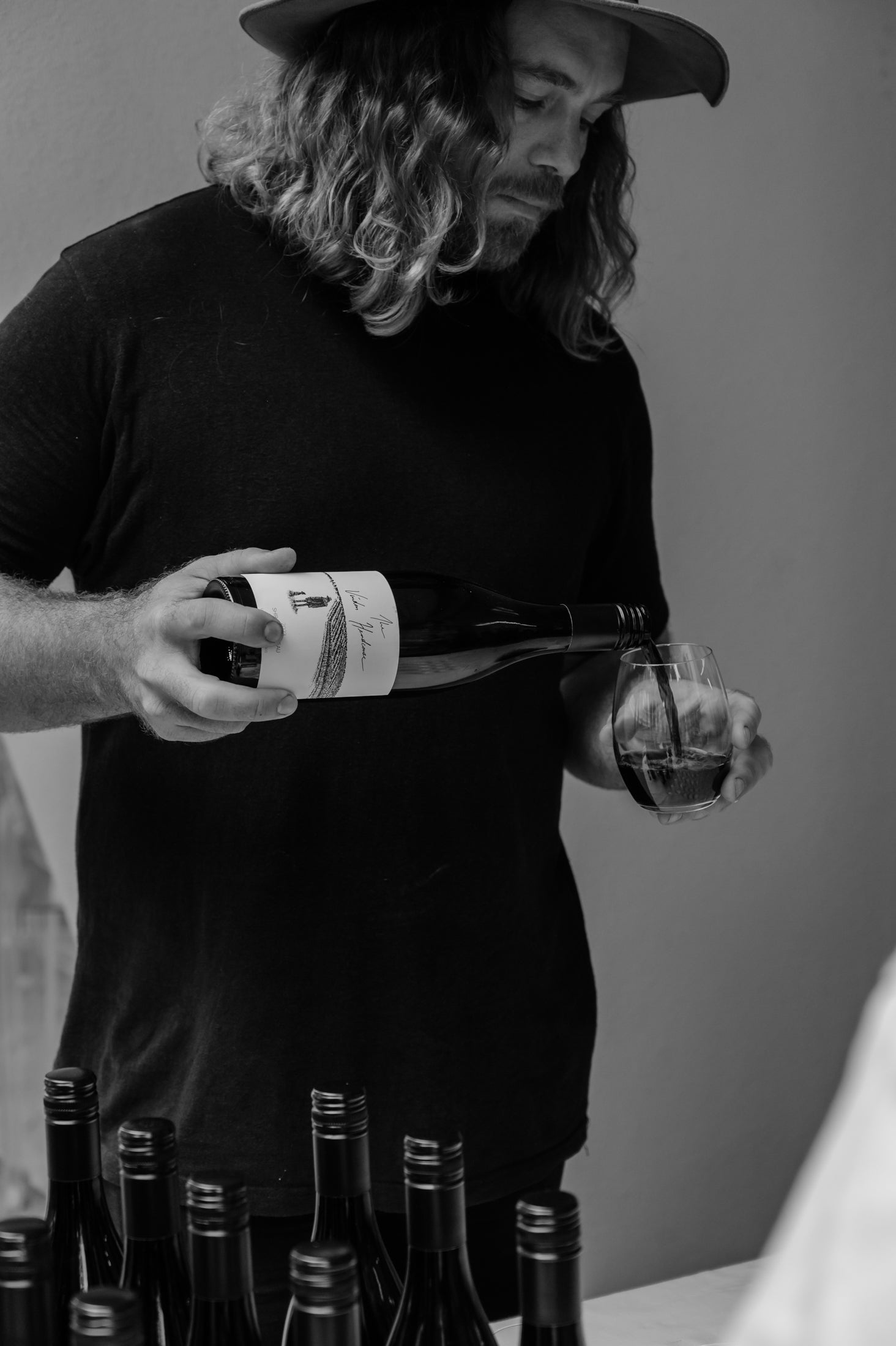 Man with long hair wearing a hat pouring red wine into a glass, surrounded by wine bottles on a table. Black and white image.