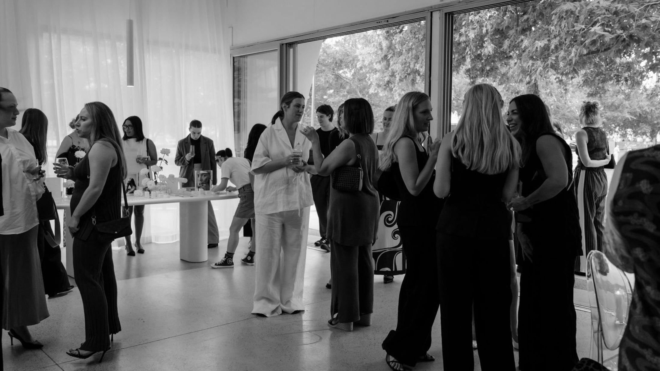 Black-and-white image of a luxury jewellery event, guests mingling indoors, elegant attire, large windows, and a display table with items.